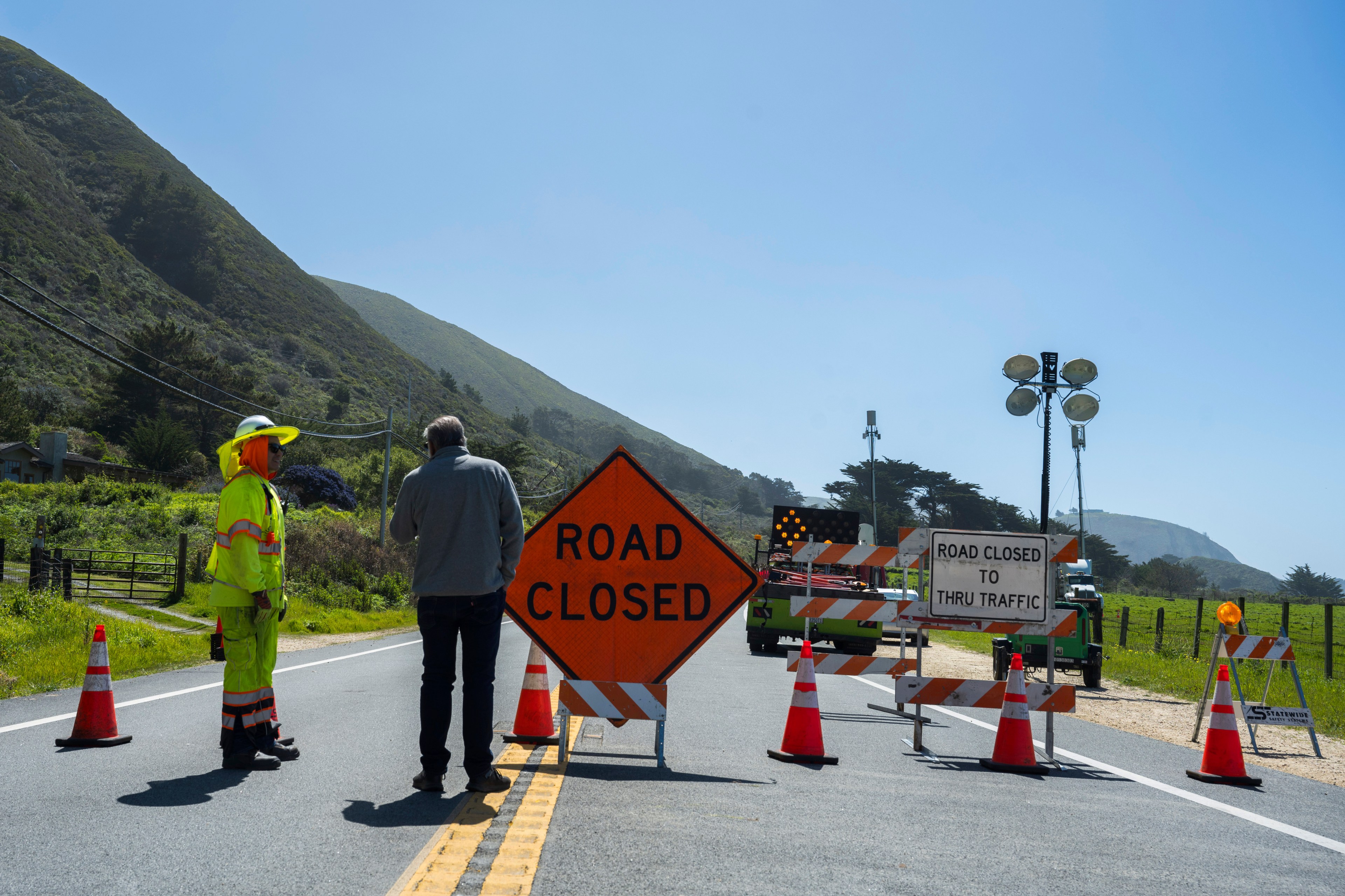 A road worker in hi-vis gear and a person stand by a &quot;ROAD CLOSED&quot; sign amidst traffic cones and barriers, with a green hill in the background.