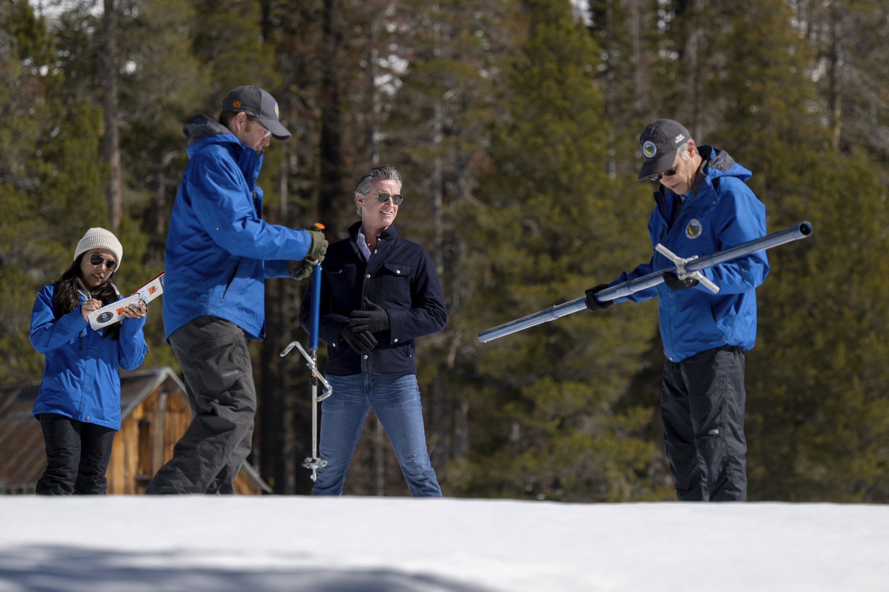 Four people in winter wear stand on snow, holding biathlon rifles, with trees in the background.