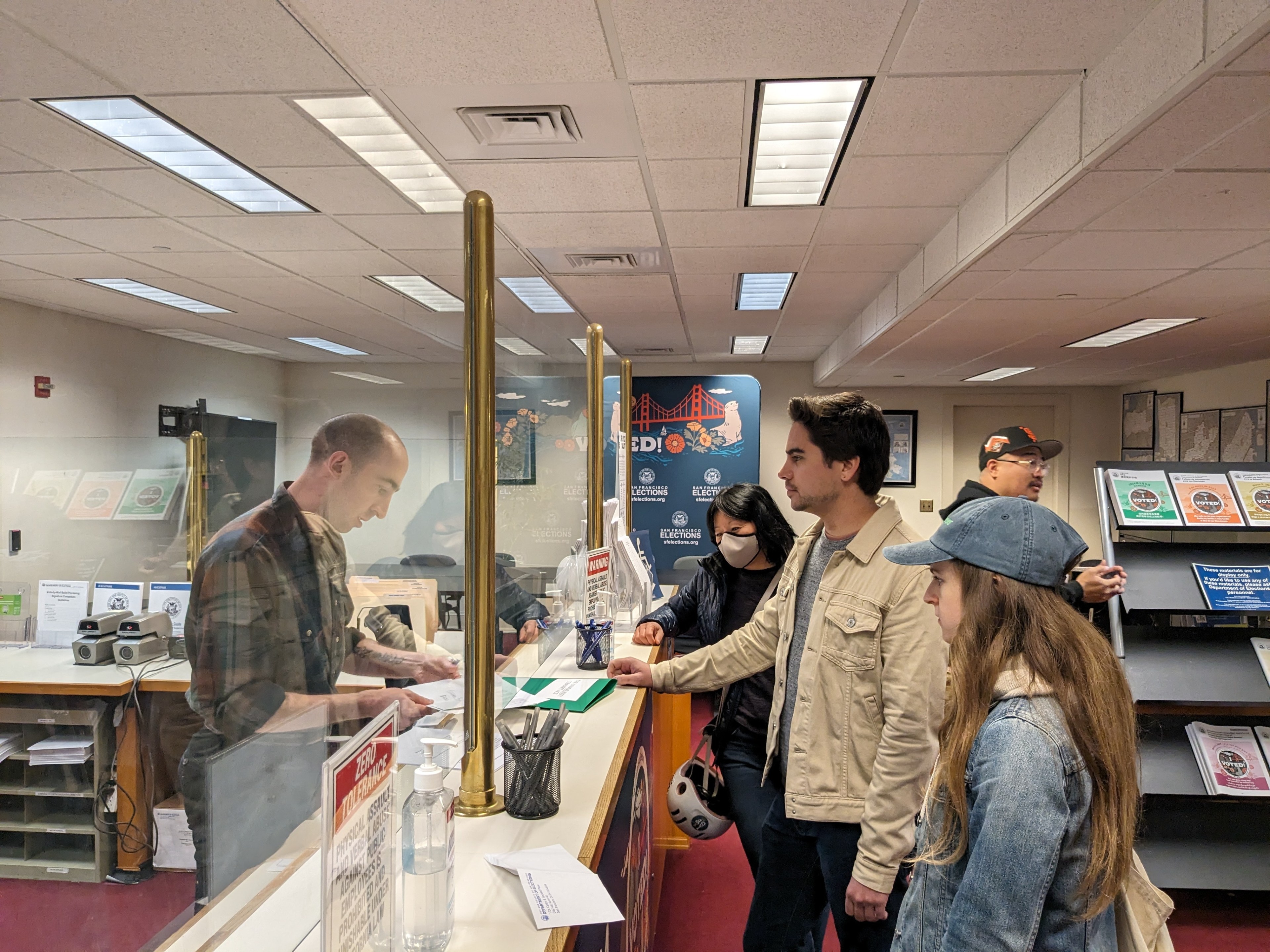People queuing at a post office counter, some are wearing masks, and promotional posters are visible.