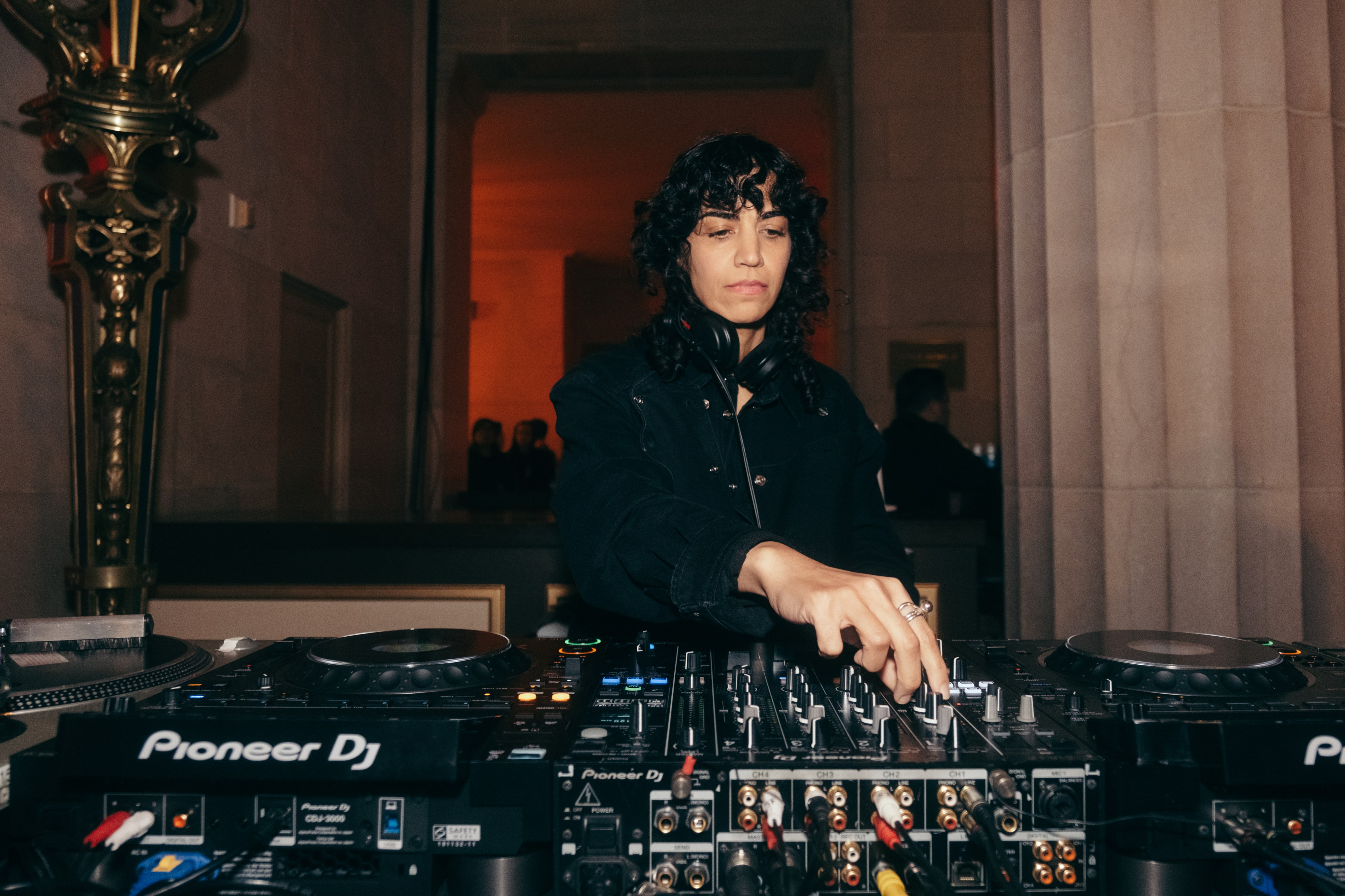 A DJ with curly hair mixes tracks on a Pioneer setup in a warmly lit event space.