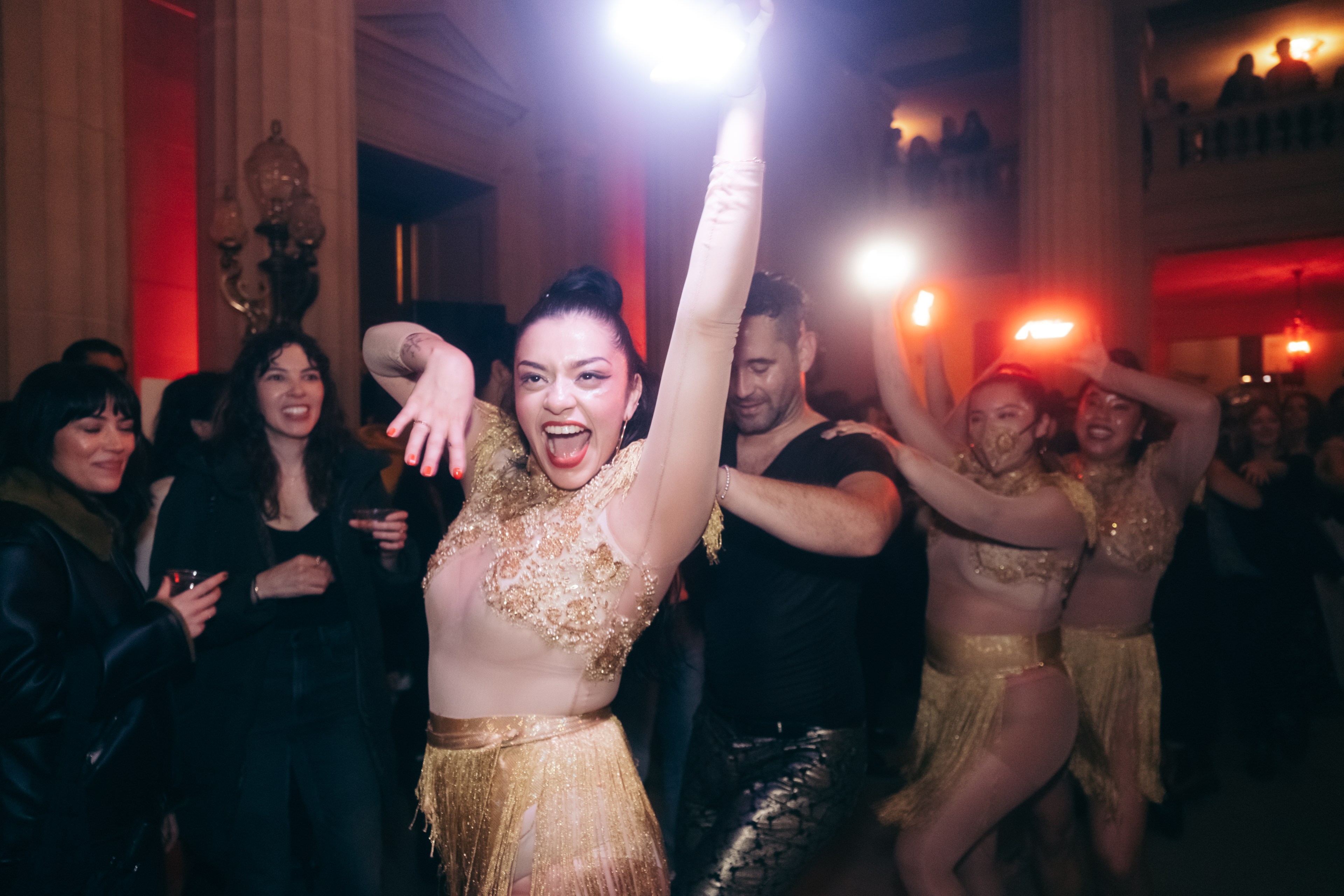 A joyous woman dances in a golden outfit at a lively party, with onlookers smiling and others dancing behind her.