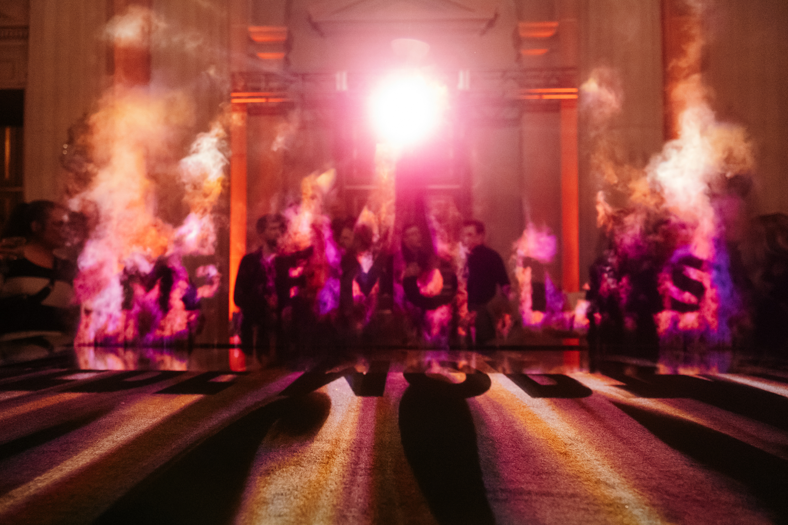A group of people behind colorful smoke plumes with a bright light source behind them, casting long shadows.