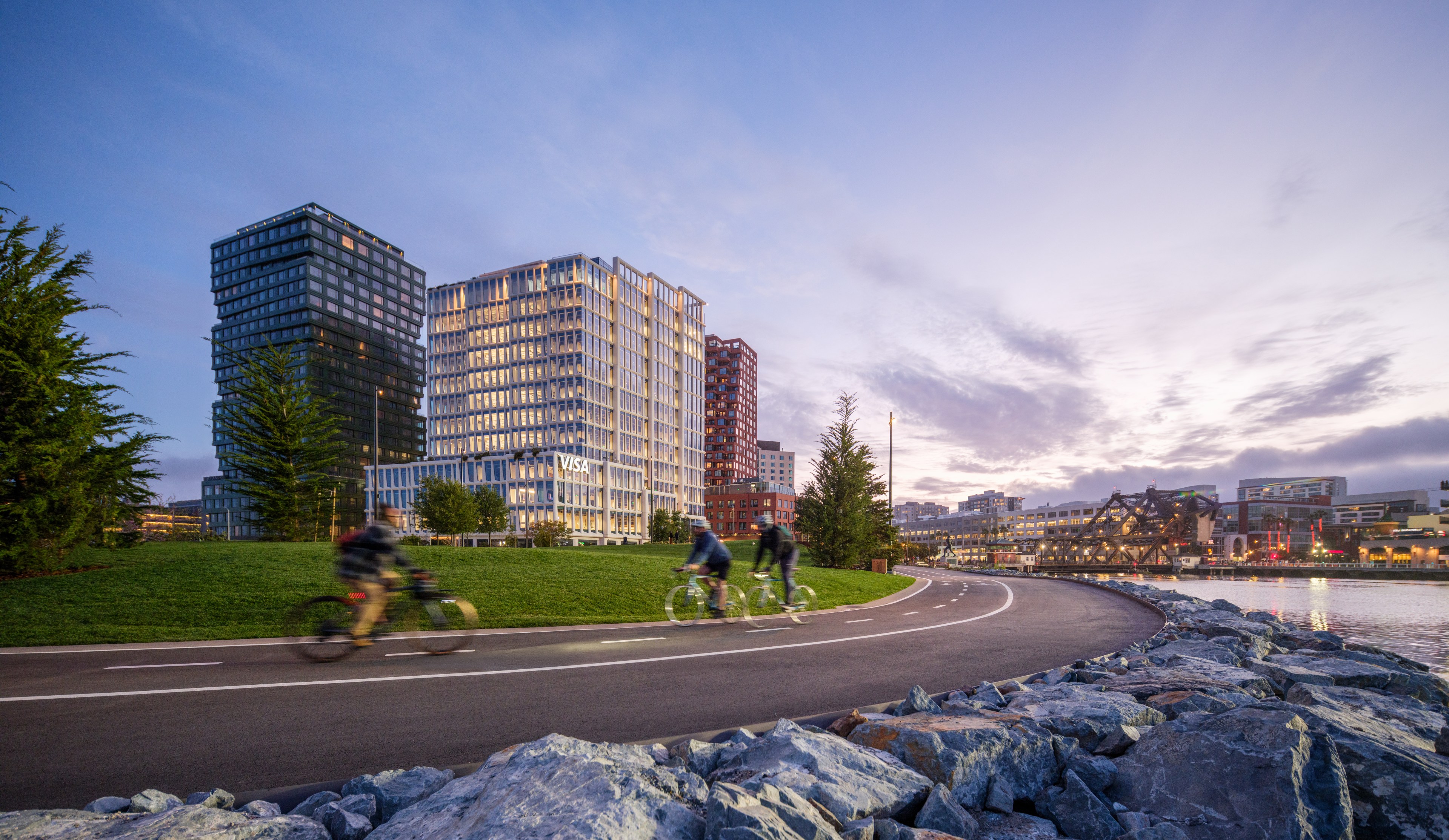 Seaside path with cyclists, modern buildings, and a sunset sky.