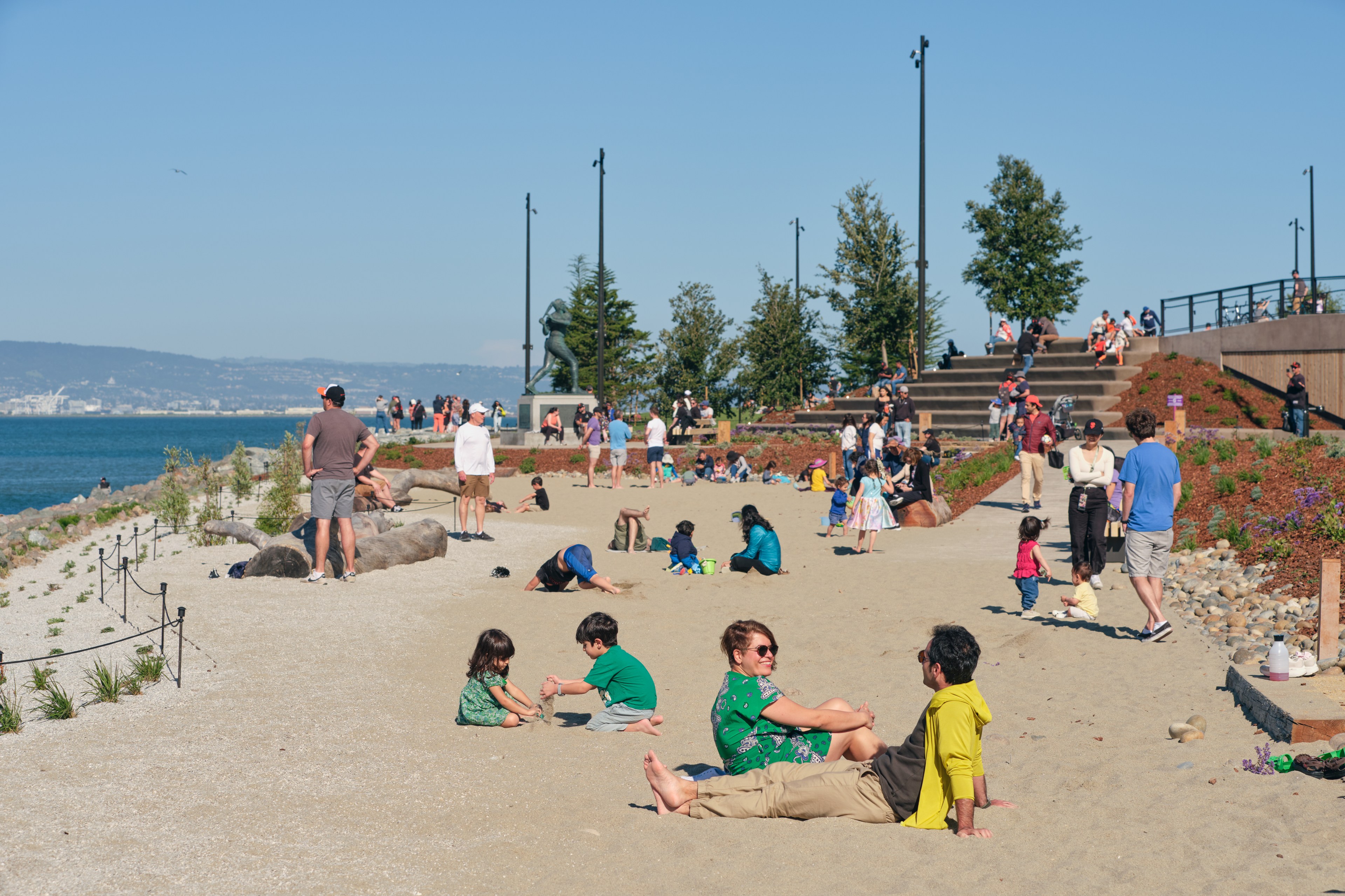 People enjoying a sunny day at a beachside park with steps, a sandy area, and a clear sky.