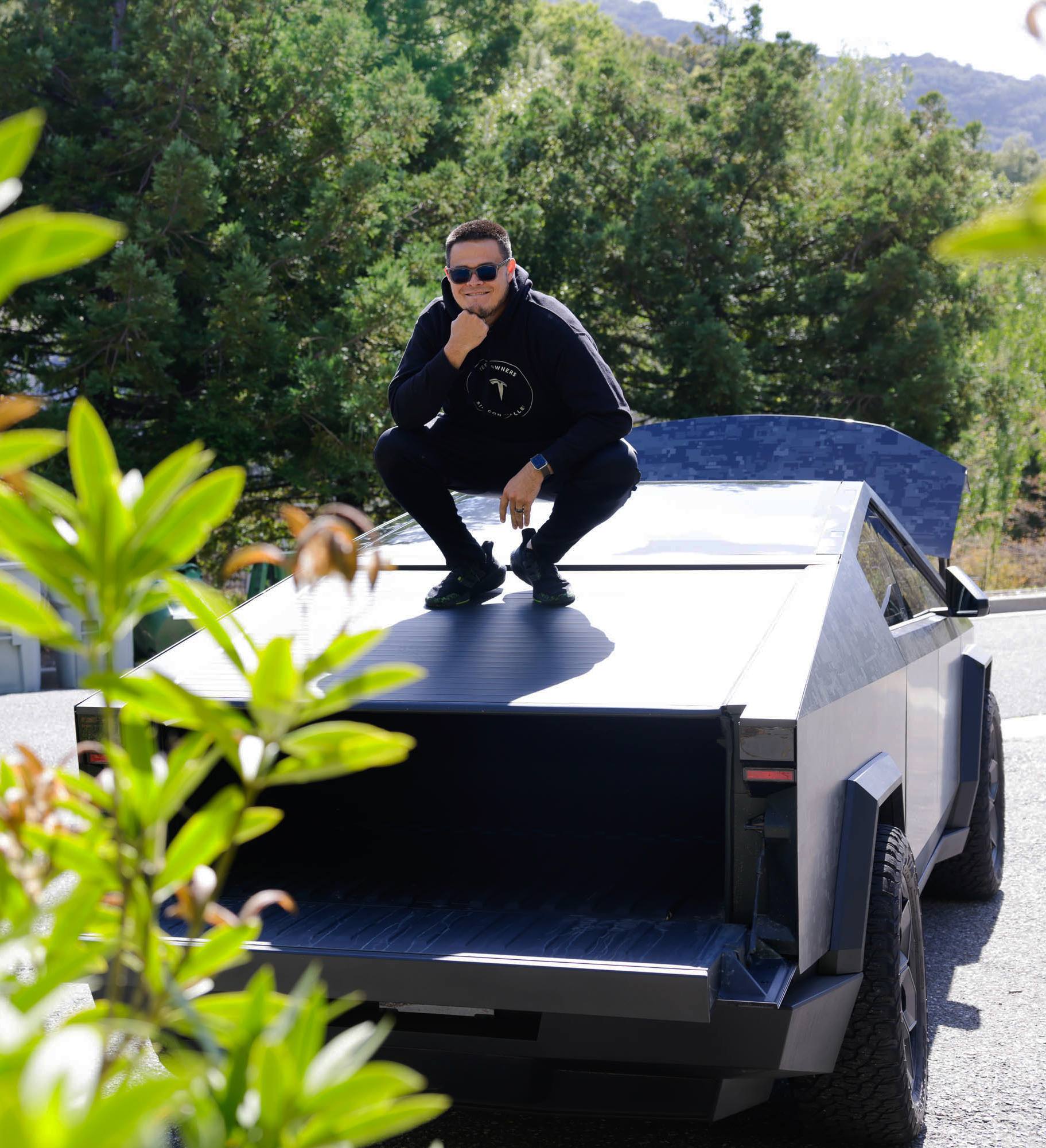 A person is squatting on top of a futuristic silver truck, wearing a black outfit and sunglasses, surrounded by greenery.