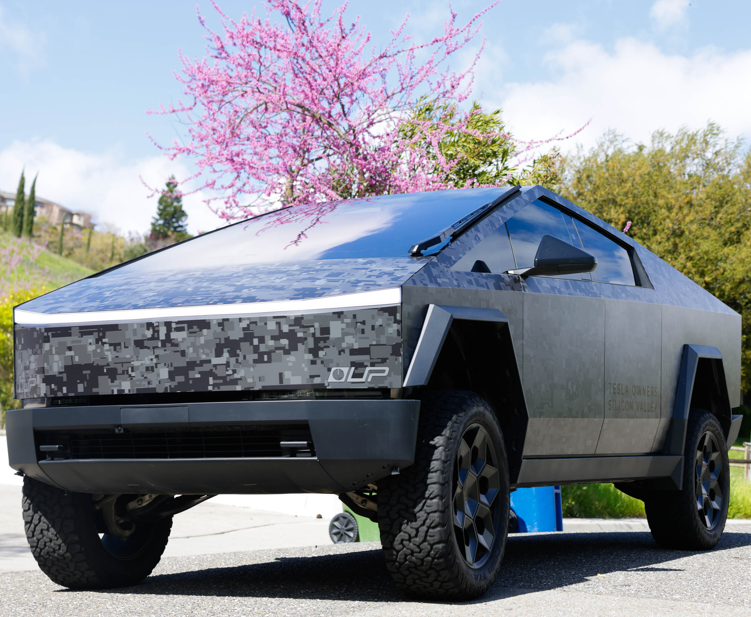A futuristic-looking electric pickup truck with a sharp, angular design parked outdoors with blooming pink trees in the background.