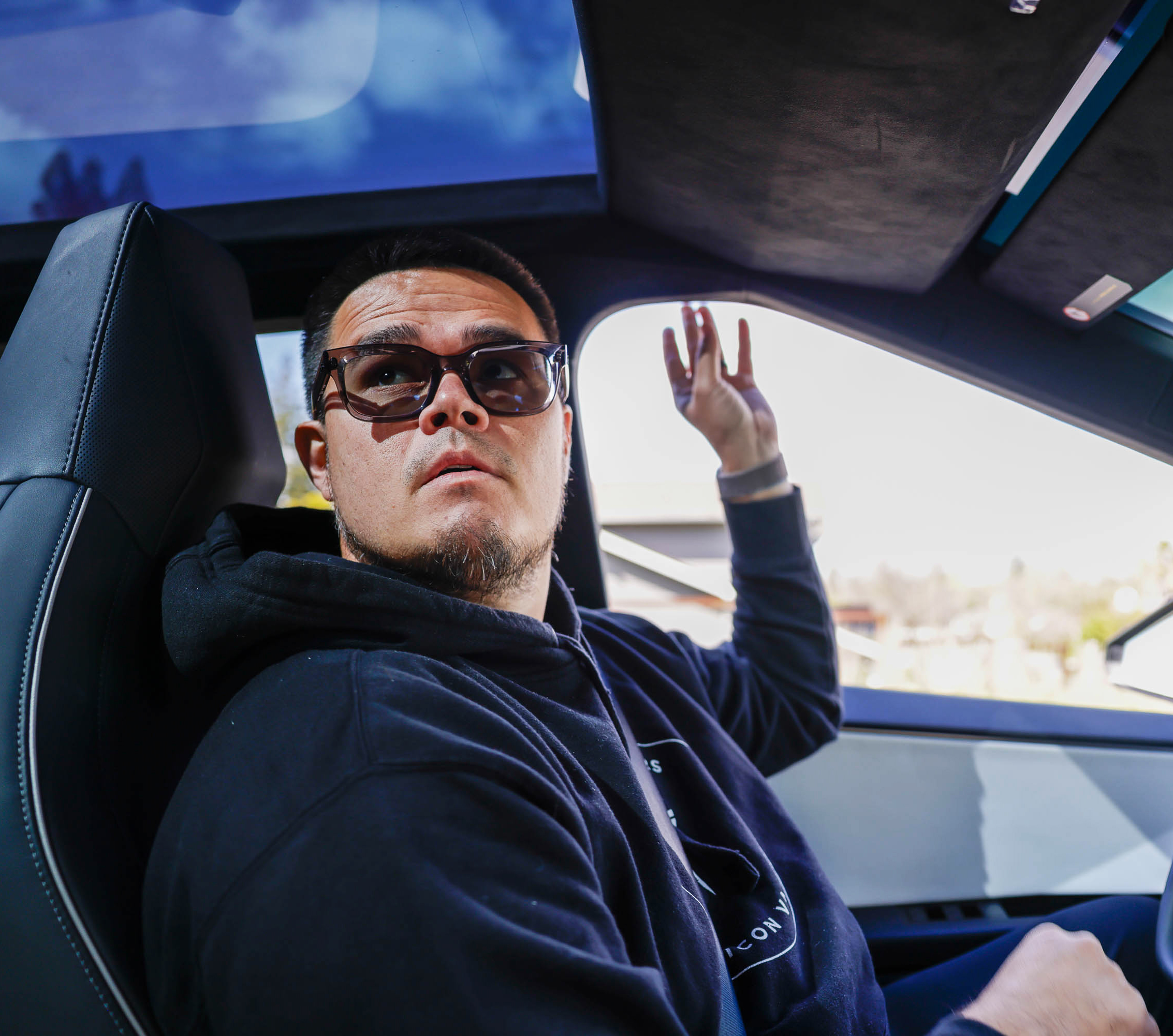 A man in sunglasses gestures inside a car with a sunroof, looking intently at the camera.
