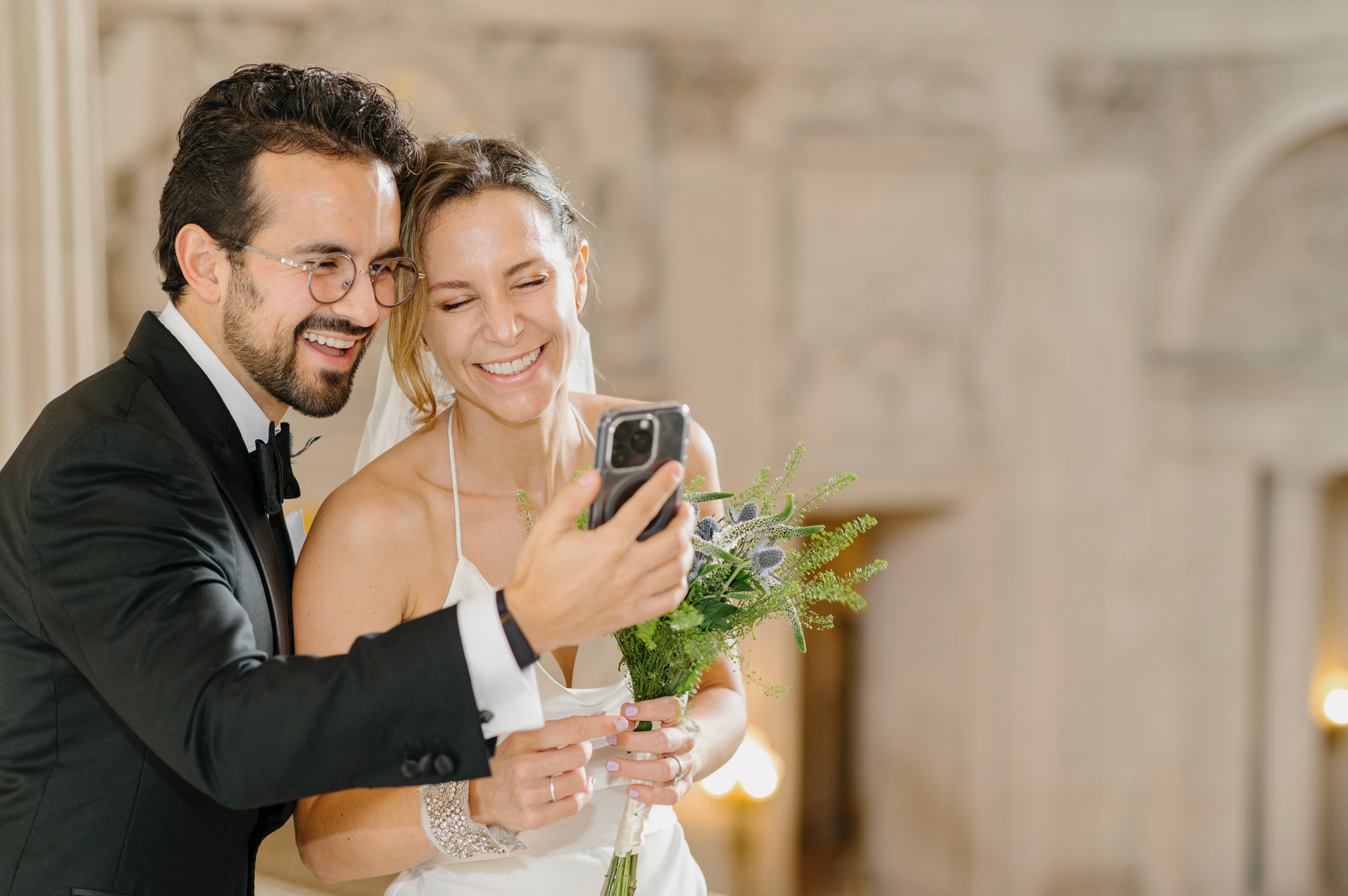 A smiling bride and groom in wedding attire share a joyful moment and look at a smartphone.