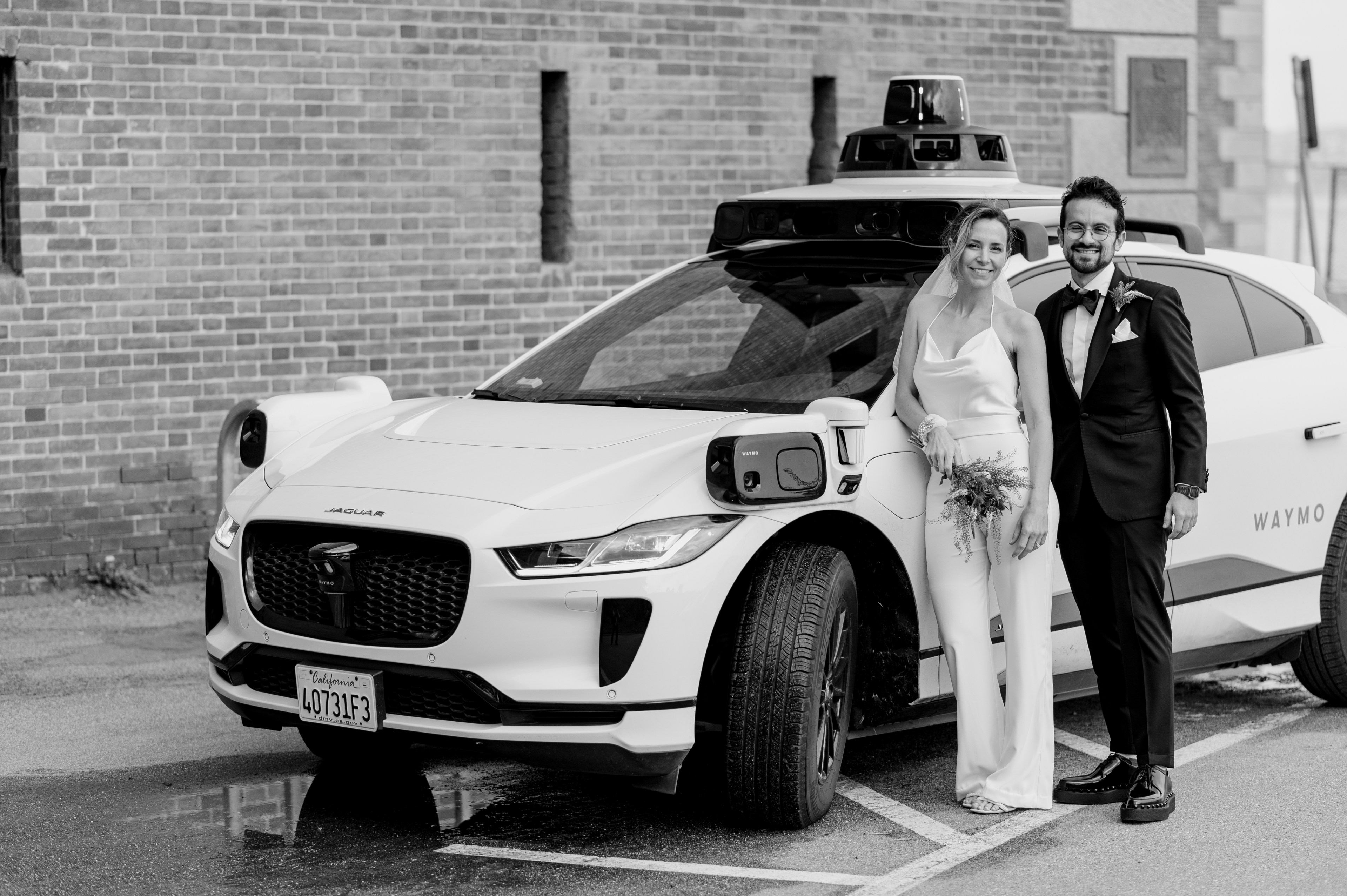 A couple in wedding attire stands by a self-driving car with sensors on the roof.