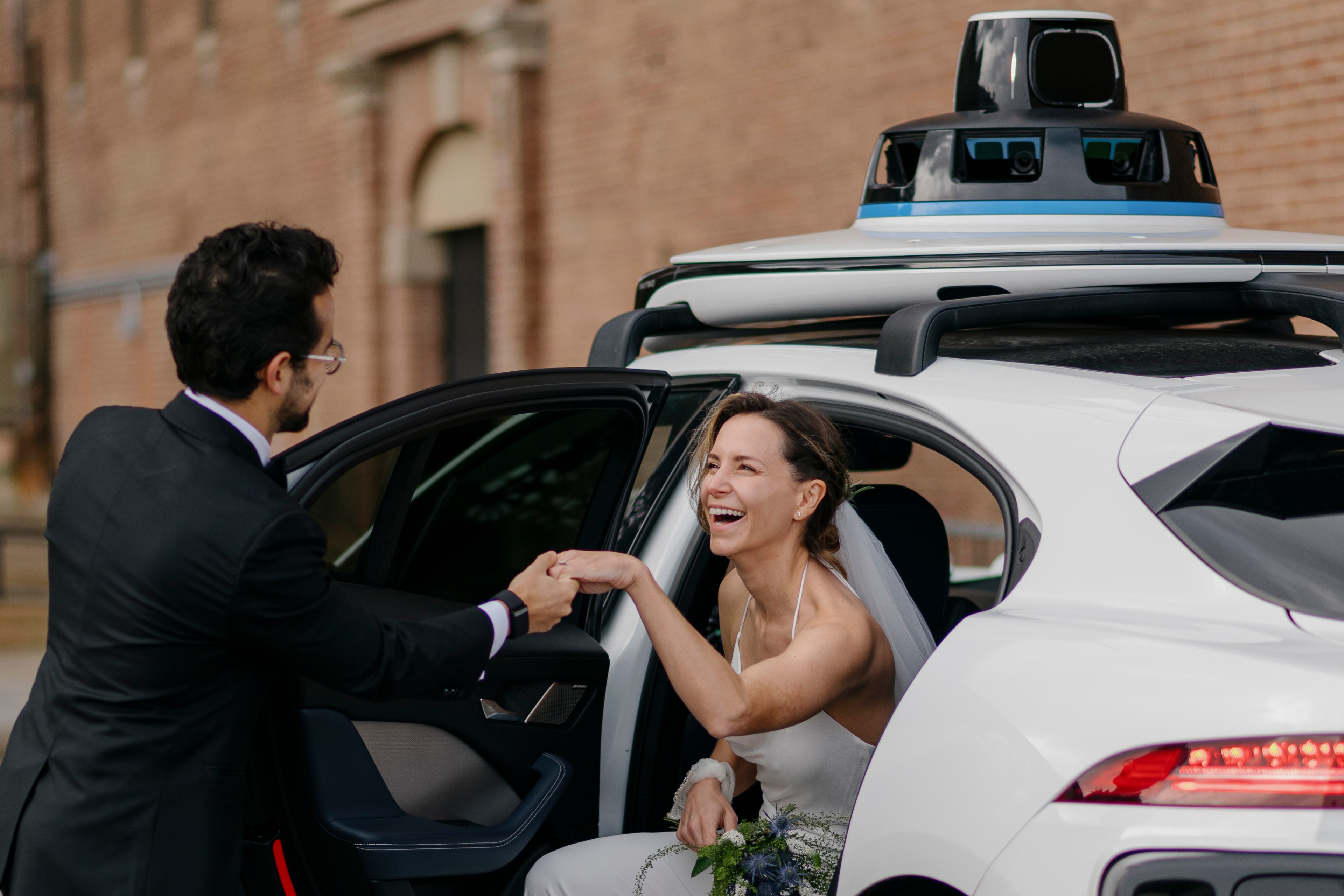 A bride in a self-driving car, with a sensor-laden roof, smiles and takes a man's hand as she gets out of the vehicle.
