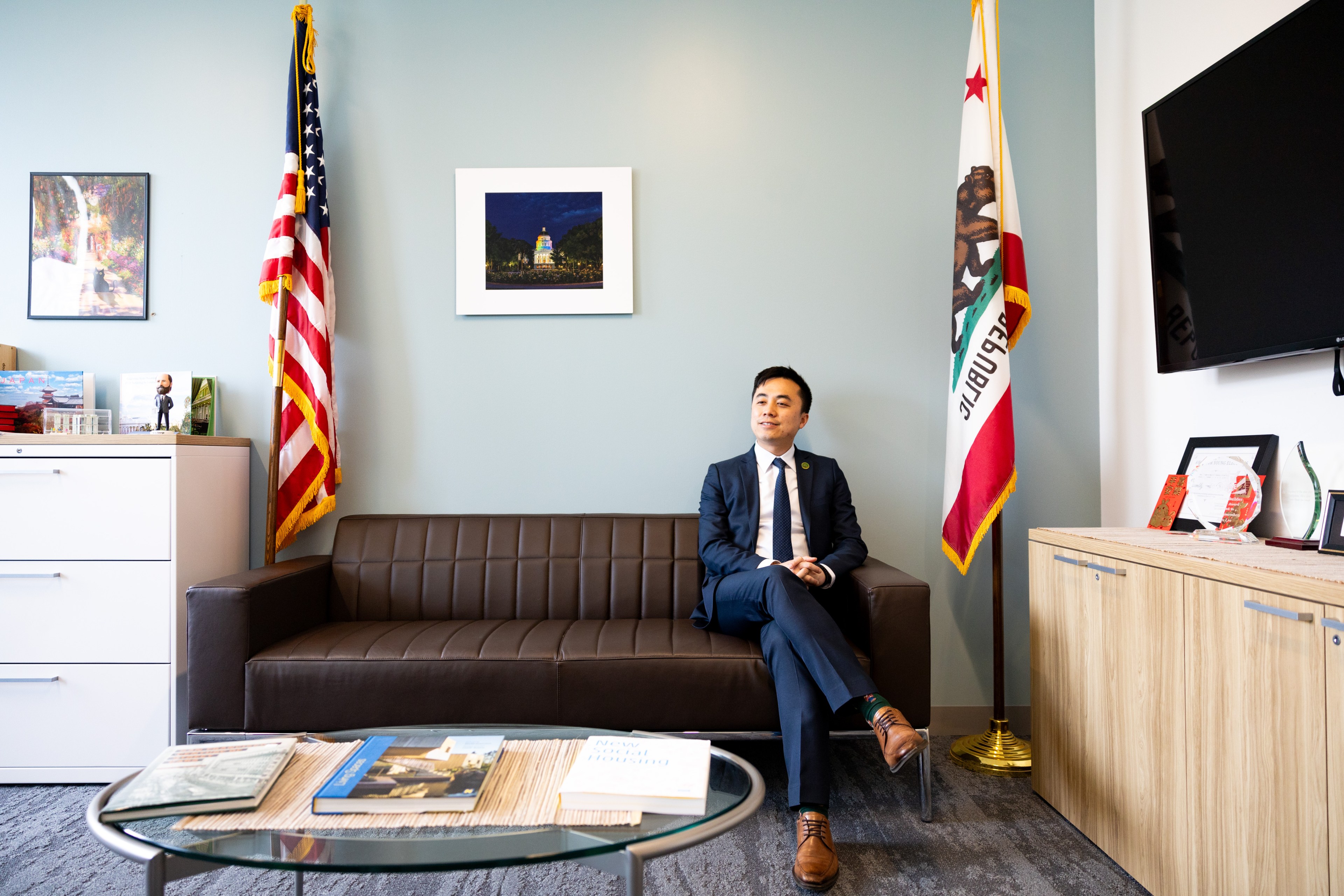 A man in a suit sits on a couch in an office with U.S. and California flags, artwork, and a TV.