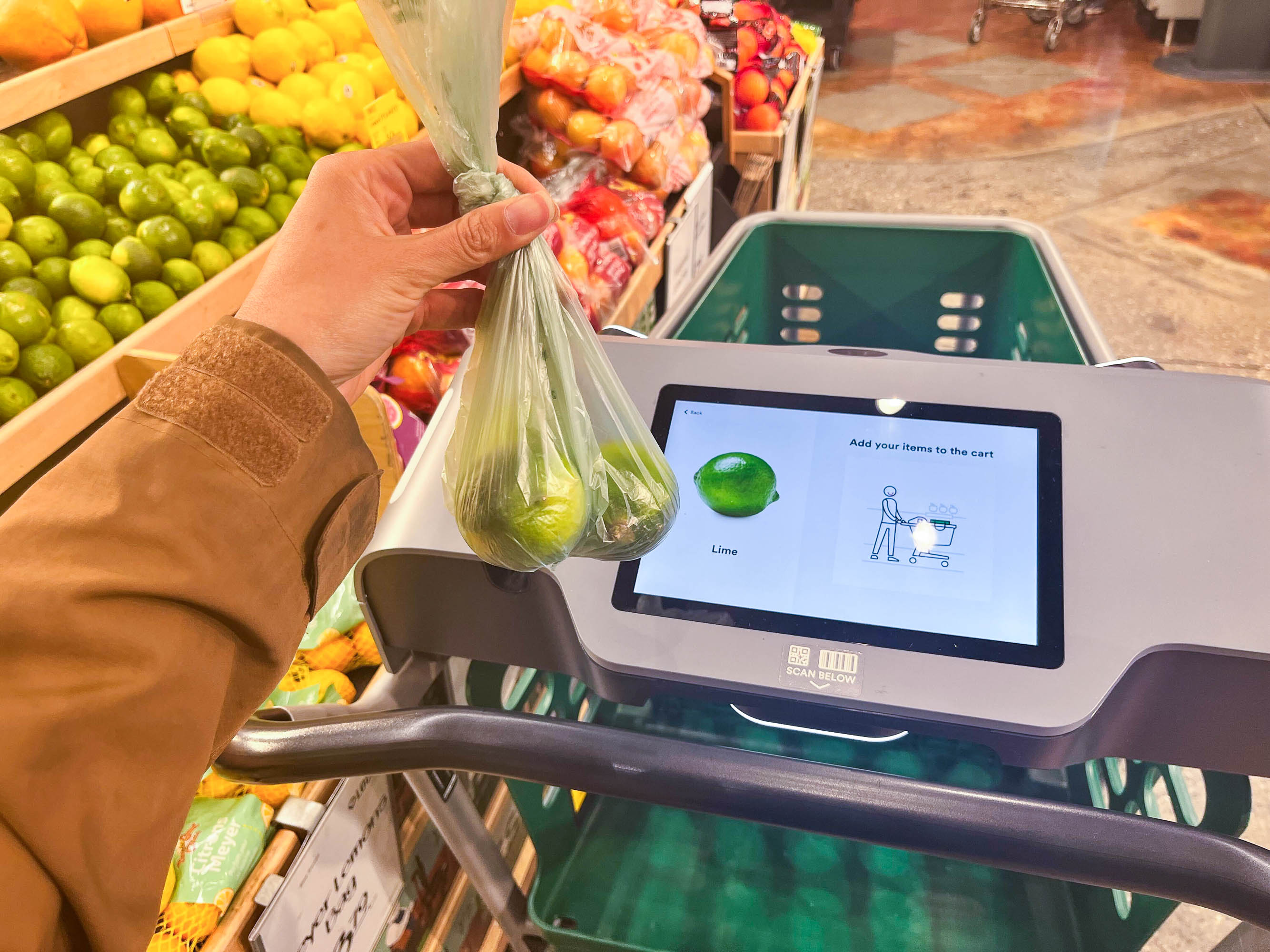 A hand holds a bag of limes over a shopping cart with a built-in screen displaying a lime image and "Add your items to the cart" text.