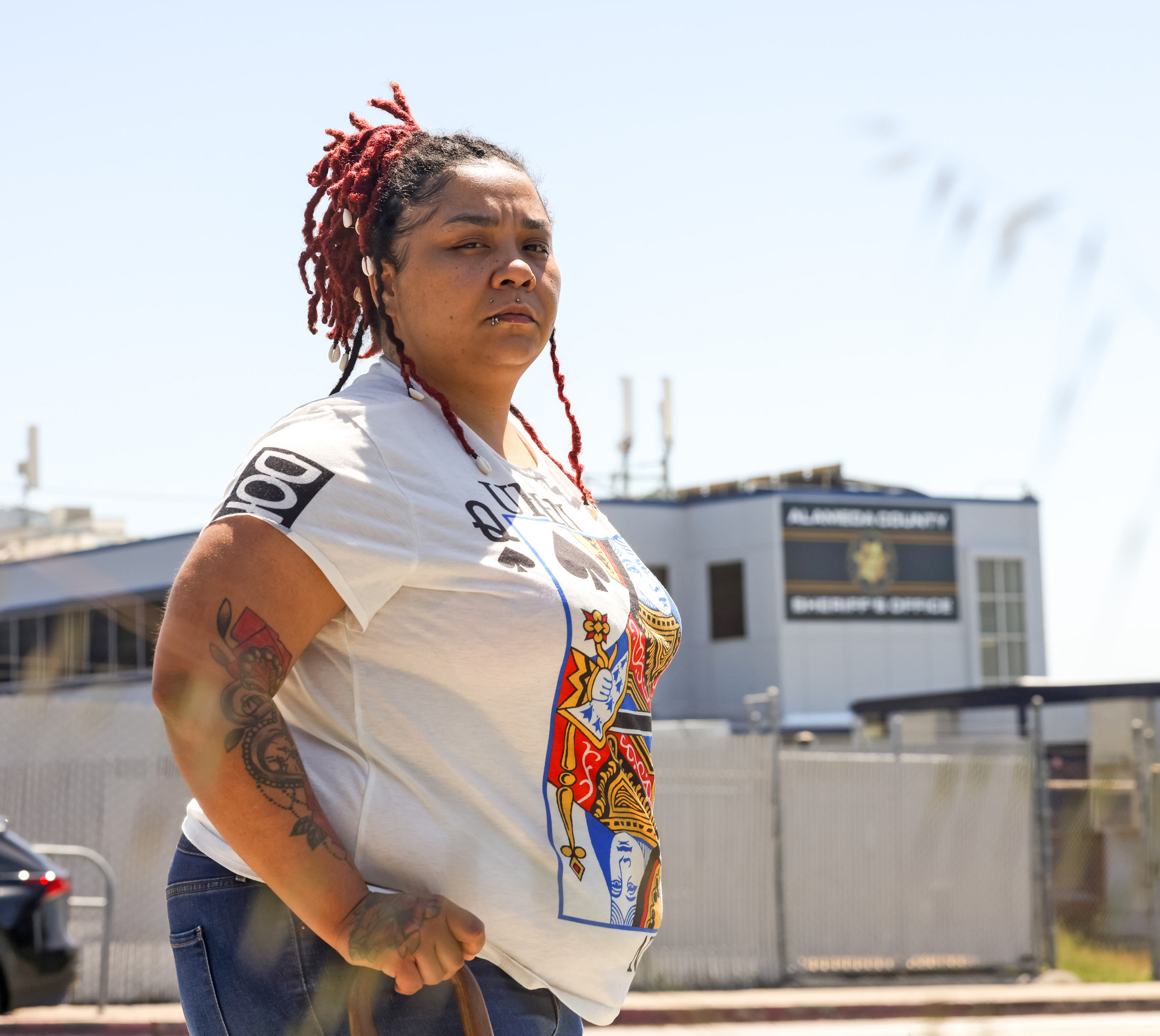 A person with red-tinged dreadlocks, tattoos, and a playing card t-shirt stands confidently outdoors.