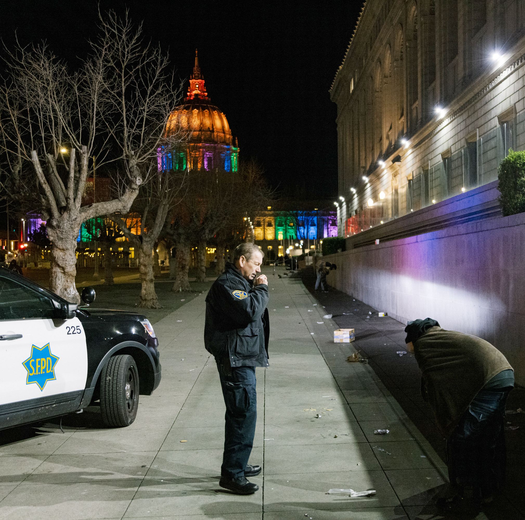 A police officer observing a person near a lit-up building at night, with a patrol car nearby.