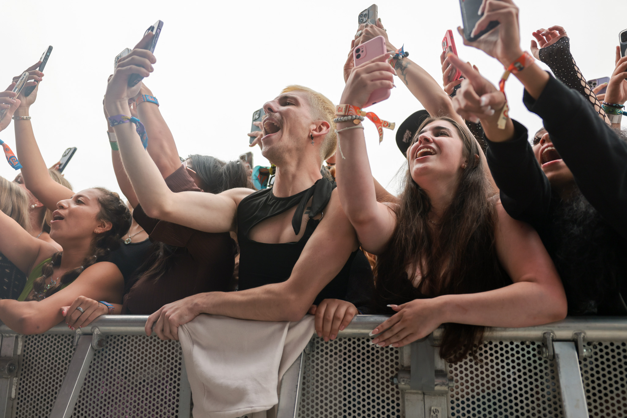 a crowd of people cheers and holds up their phones at the very front of an outdoor festival stage