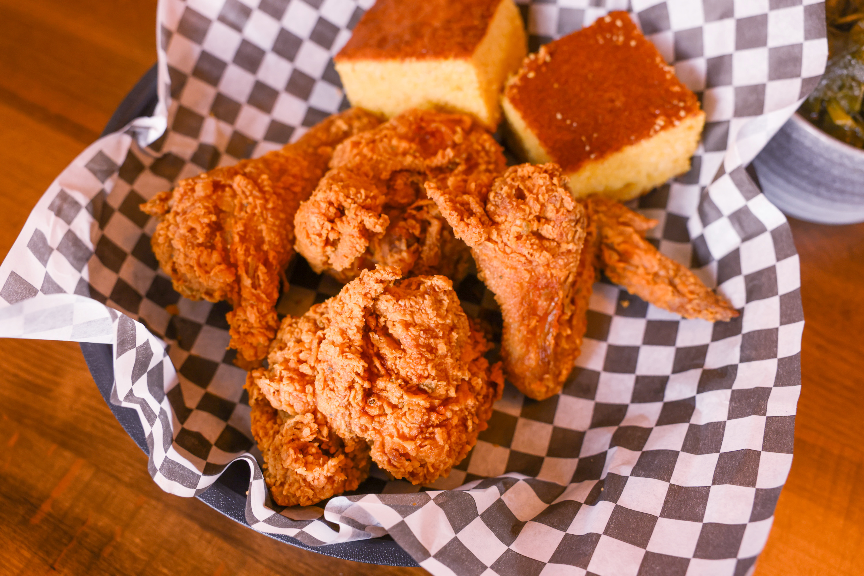 Fried chicken and cornbread in a basket with checkered paper; collard greens visible in the background.