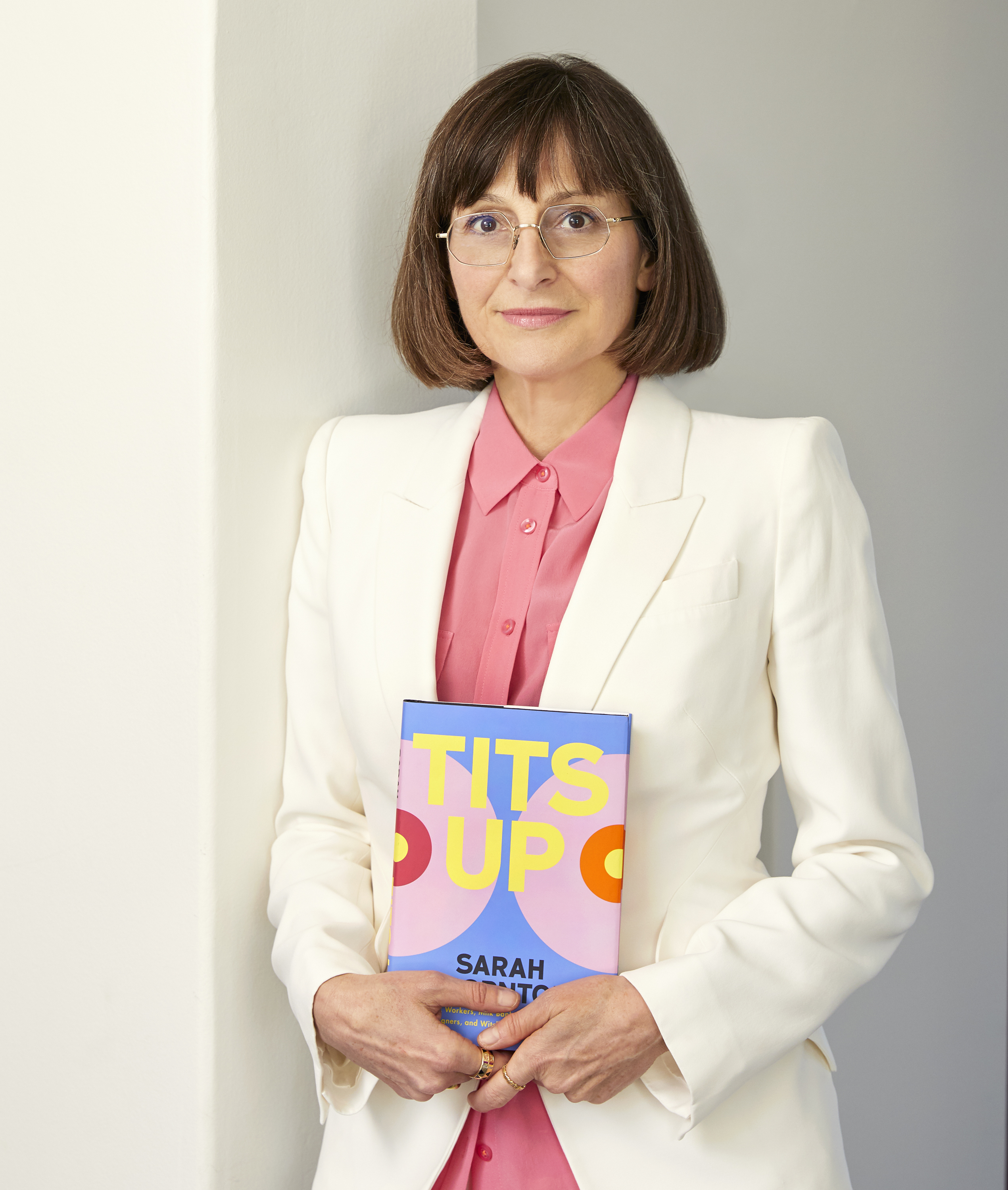 A woman in a white suit and pink shirt holding a book titled &quot;TITS UP&quot; against a plain background.