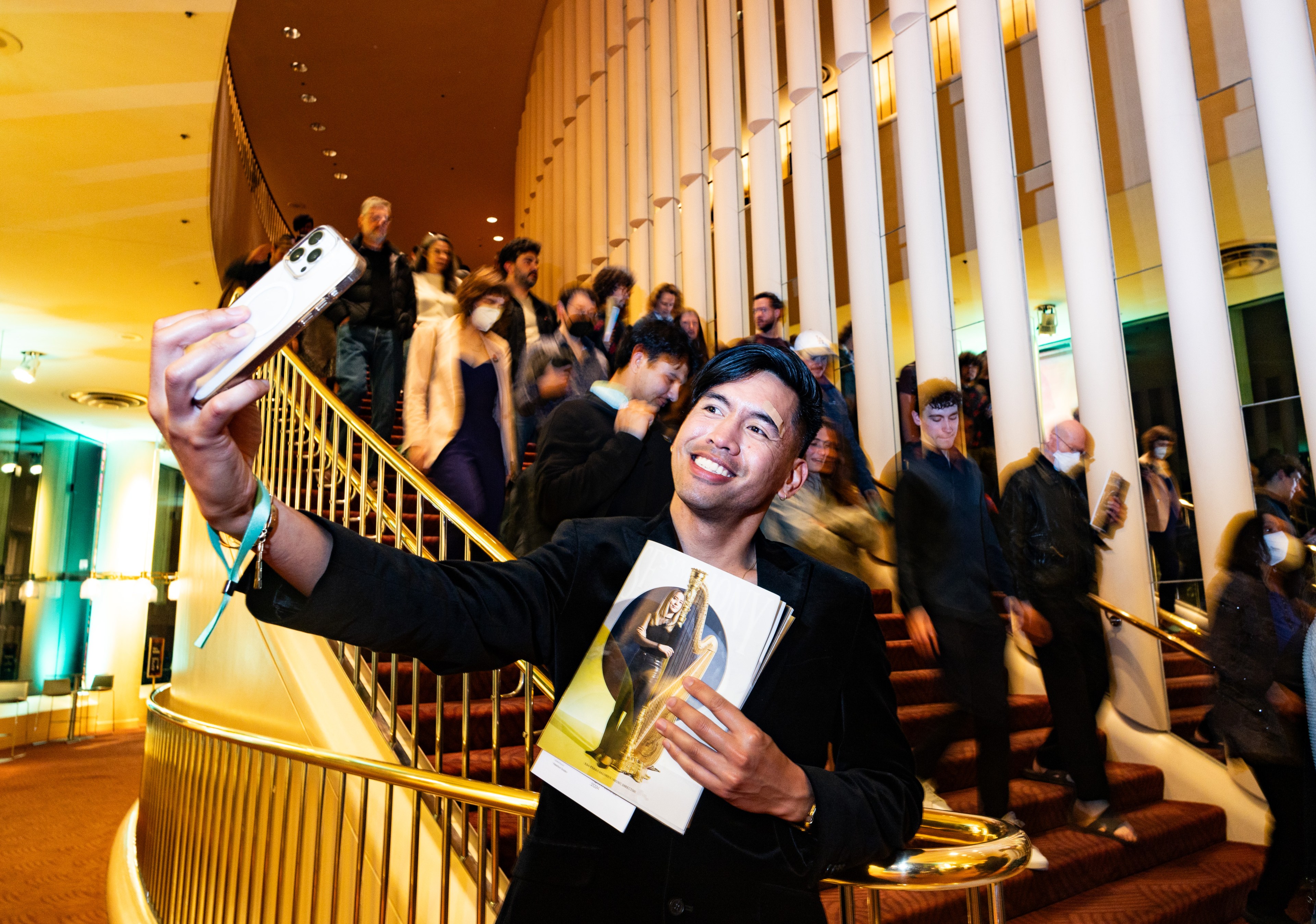 A man is taking a selfie, holding an award and a booklet, smiles on a staircase with people descending in the background.