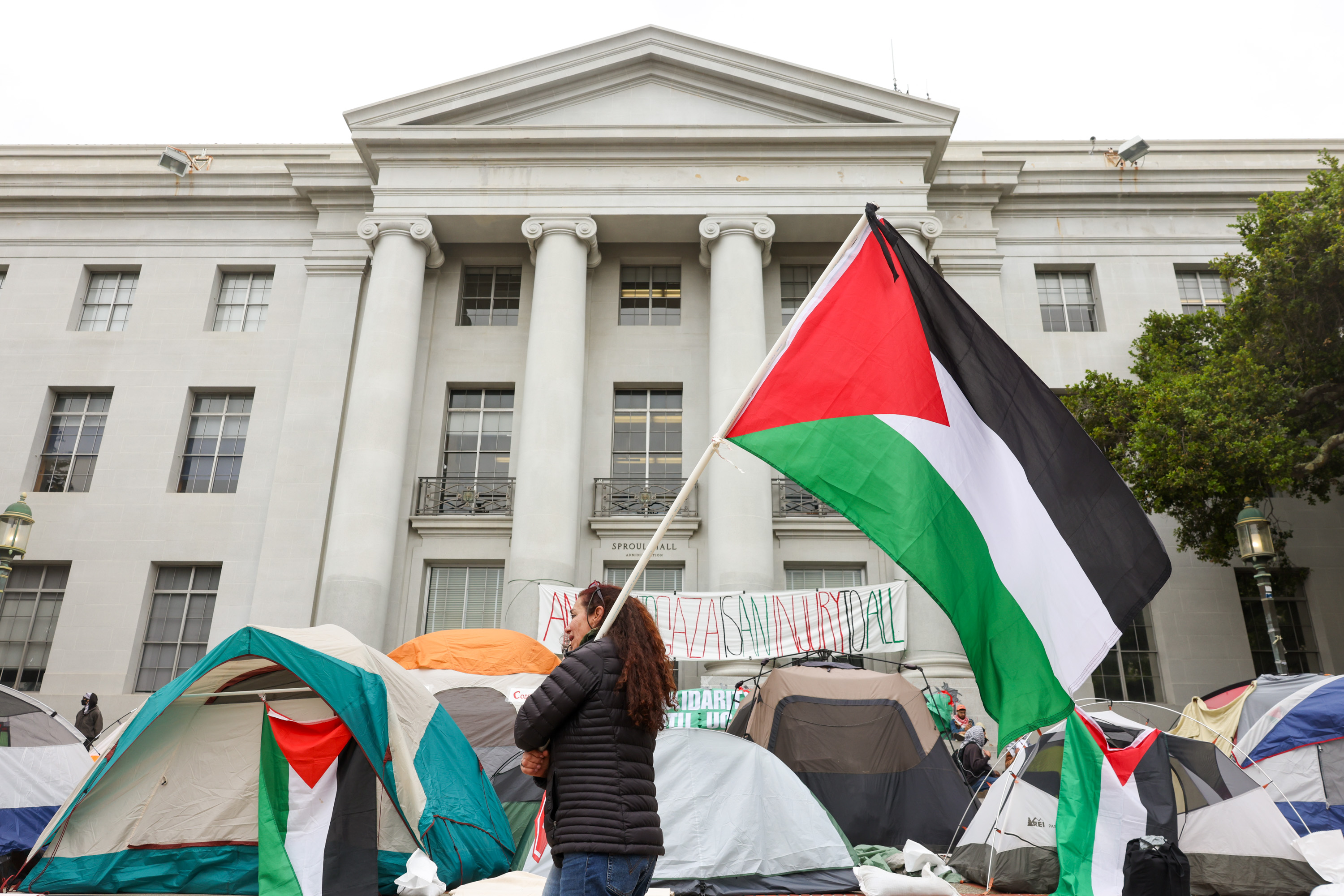 A person waves a large Palestinian flag in front of a building with tents and protest banners.