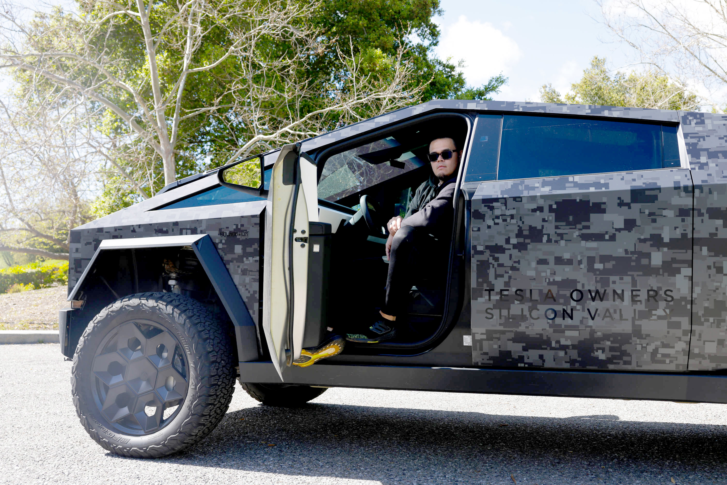 A person is sitting in a camouflaged Tesla Cybertruck with the door open, parked outdoors.