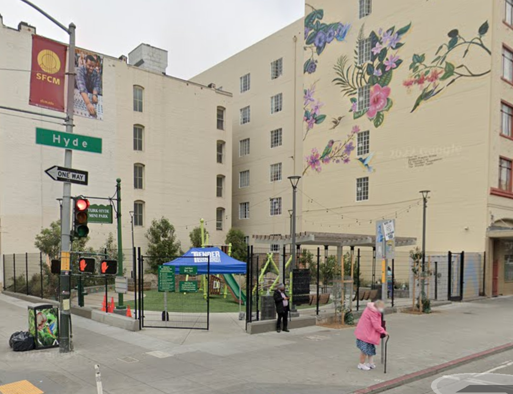 A street corner with a large mural of flowers and birds on a building, next to a small fenced park and pedestrians.