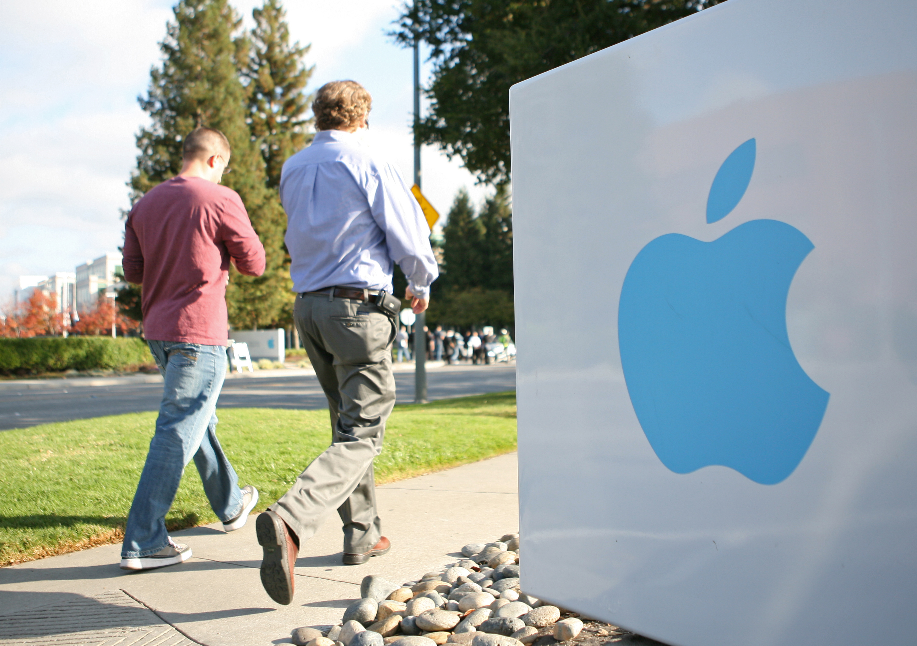 Two people are walking past a large sign with a blue Apple logo, set against a sunny outdoor background with trees.