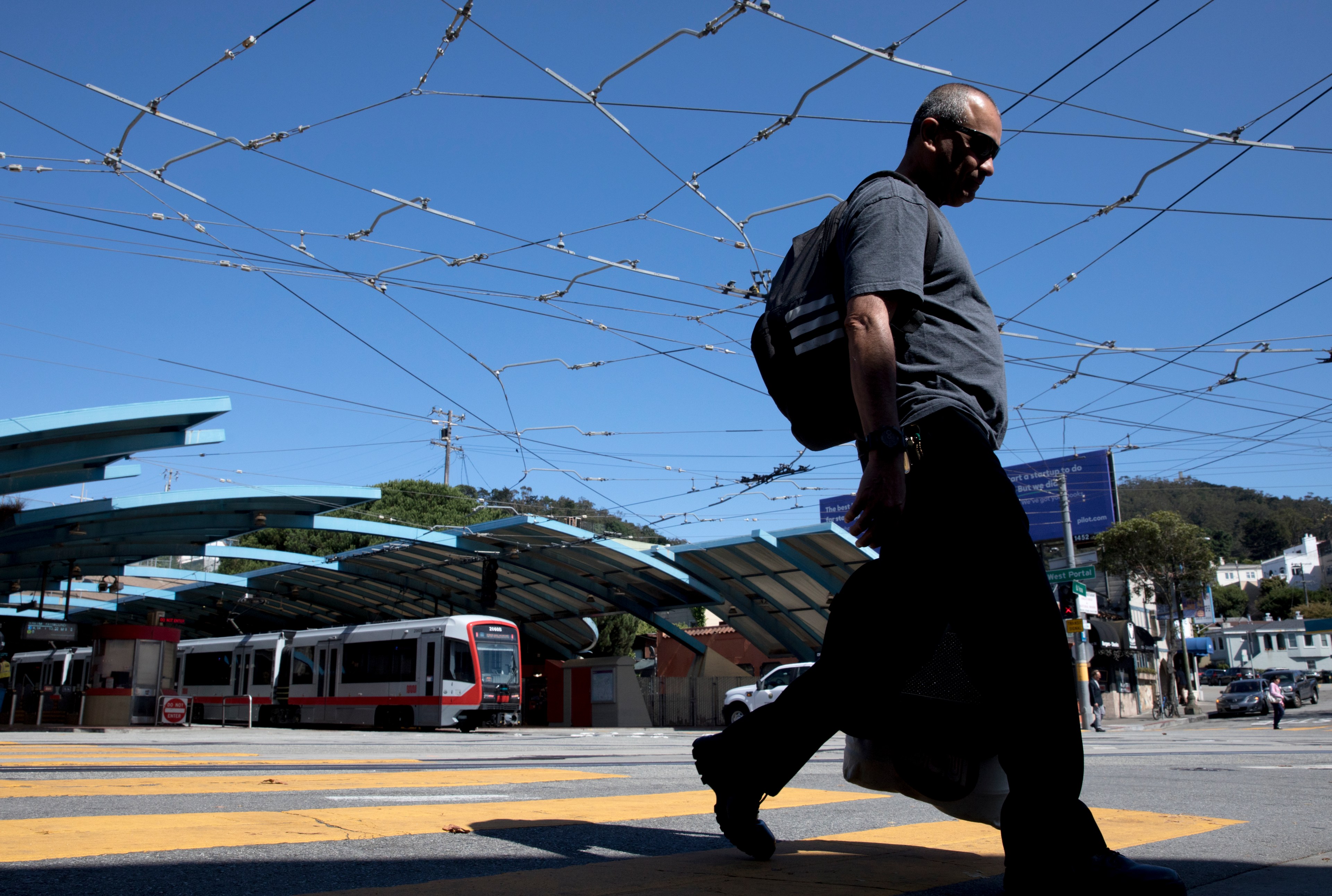 A man walks across a street with a tram station and overhead wires in the background.