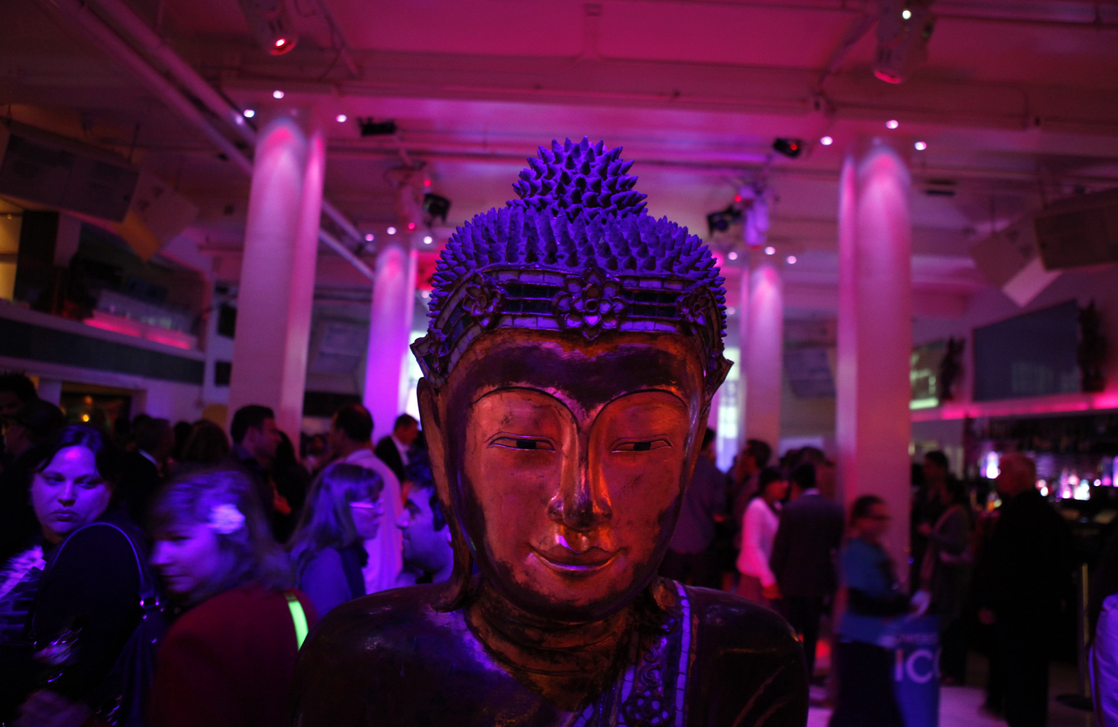 A Buddha statue is foregrounded against a lively party with purple lighting.