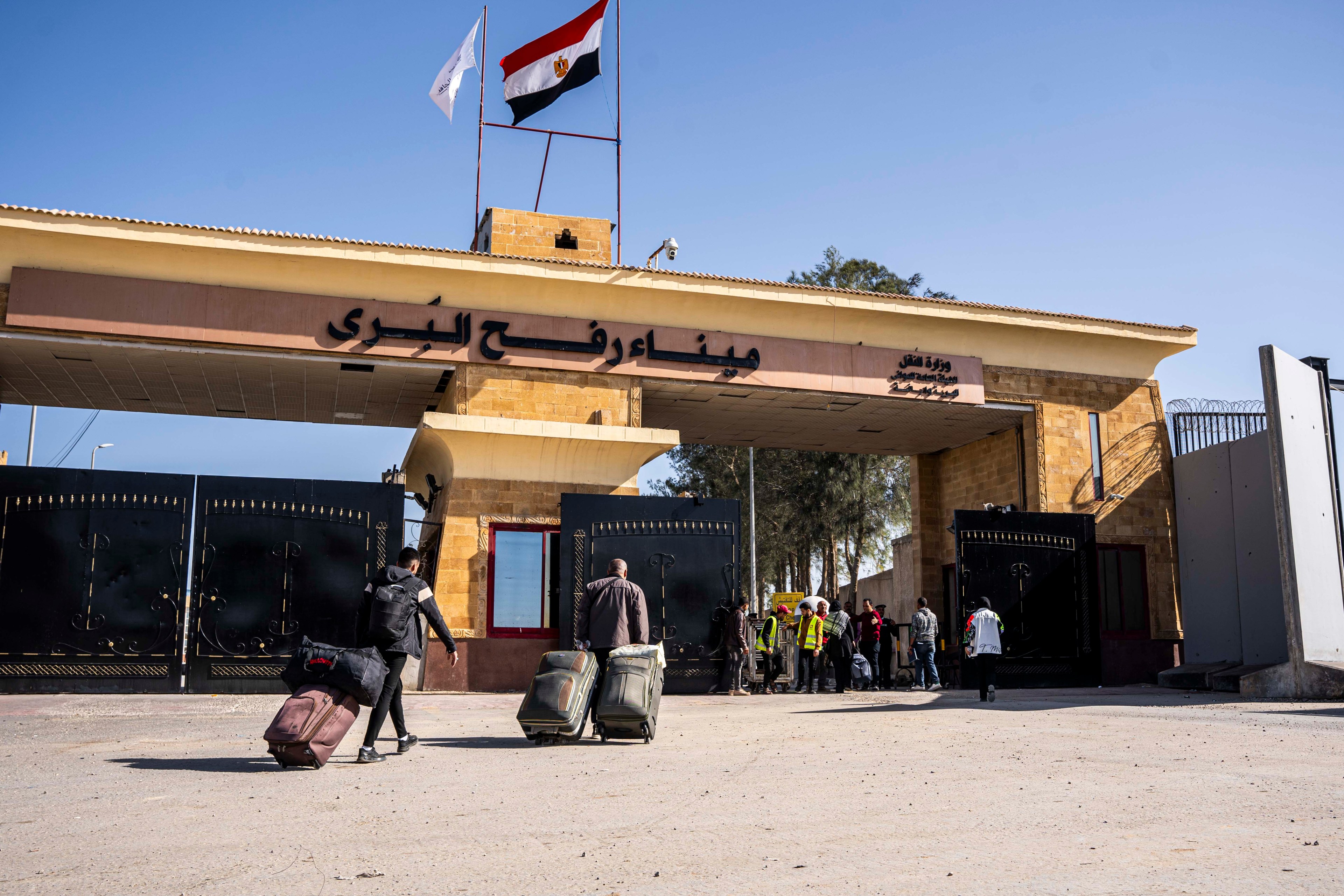 Travelers with luggage walk towards a gate beneath a building with flags and Arab script signage.