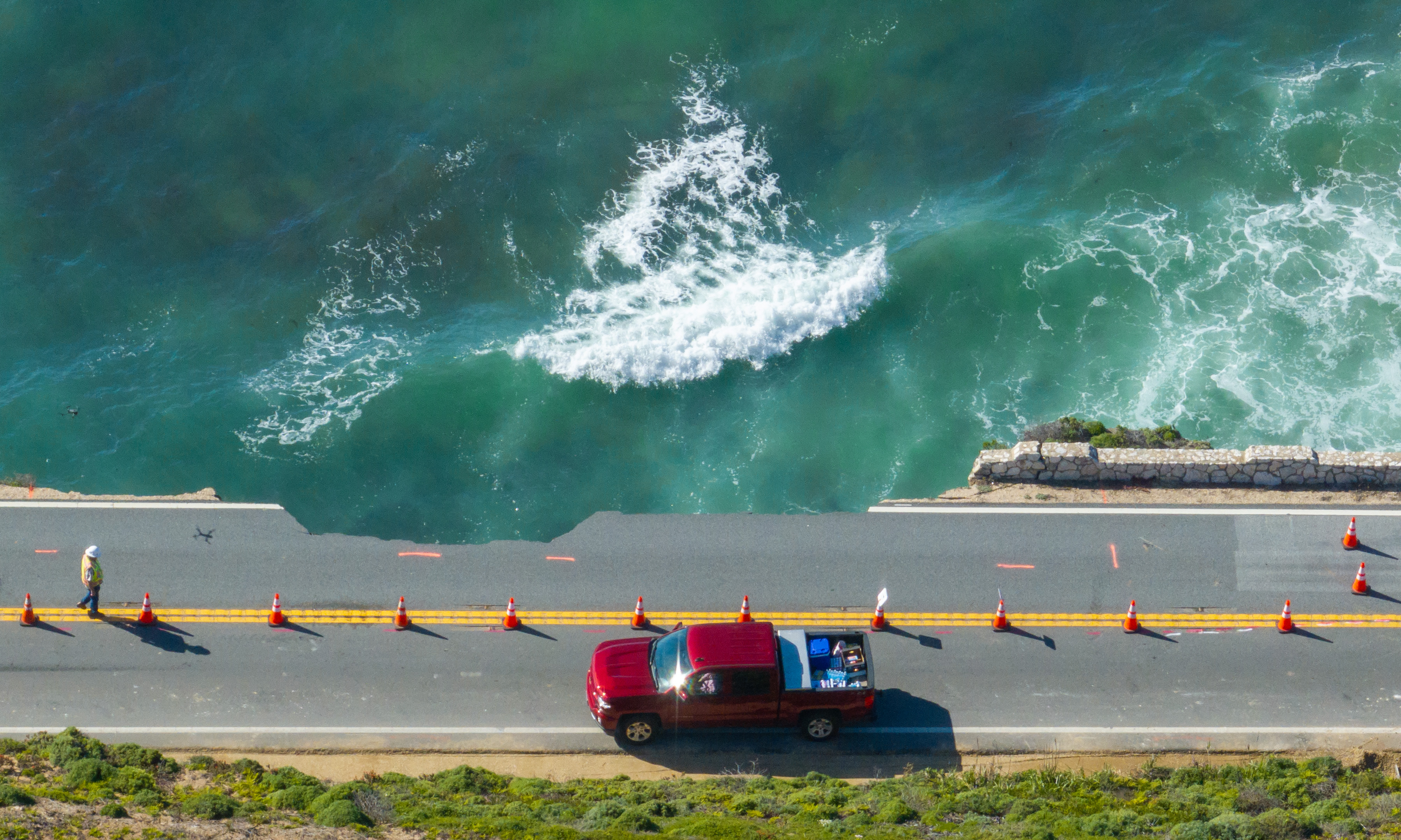 Aerial view of a red truck on a coastal road beside a turbulent teal sea and a walking worker.