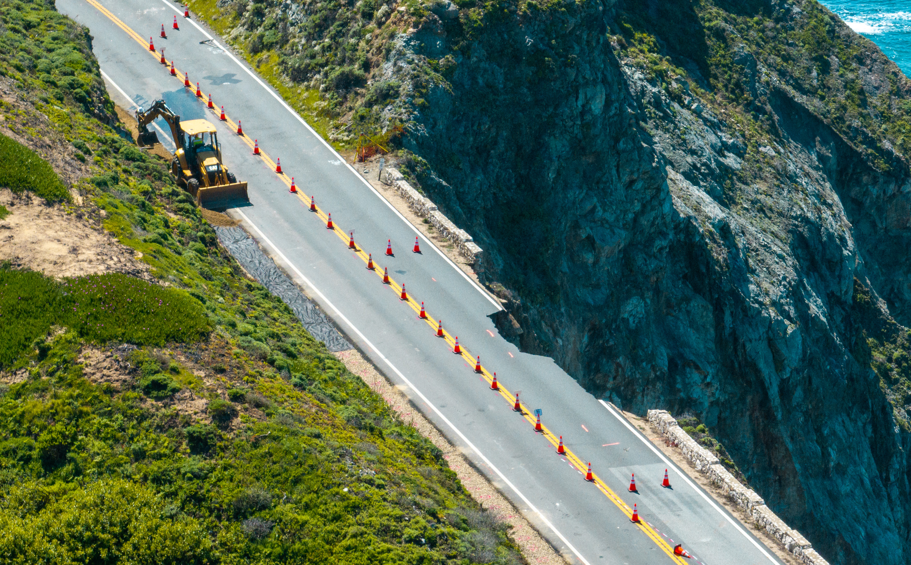 A coastal road with traffic cones and a yellow excavator working near a steep cliff overlooking a blue sea.