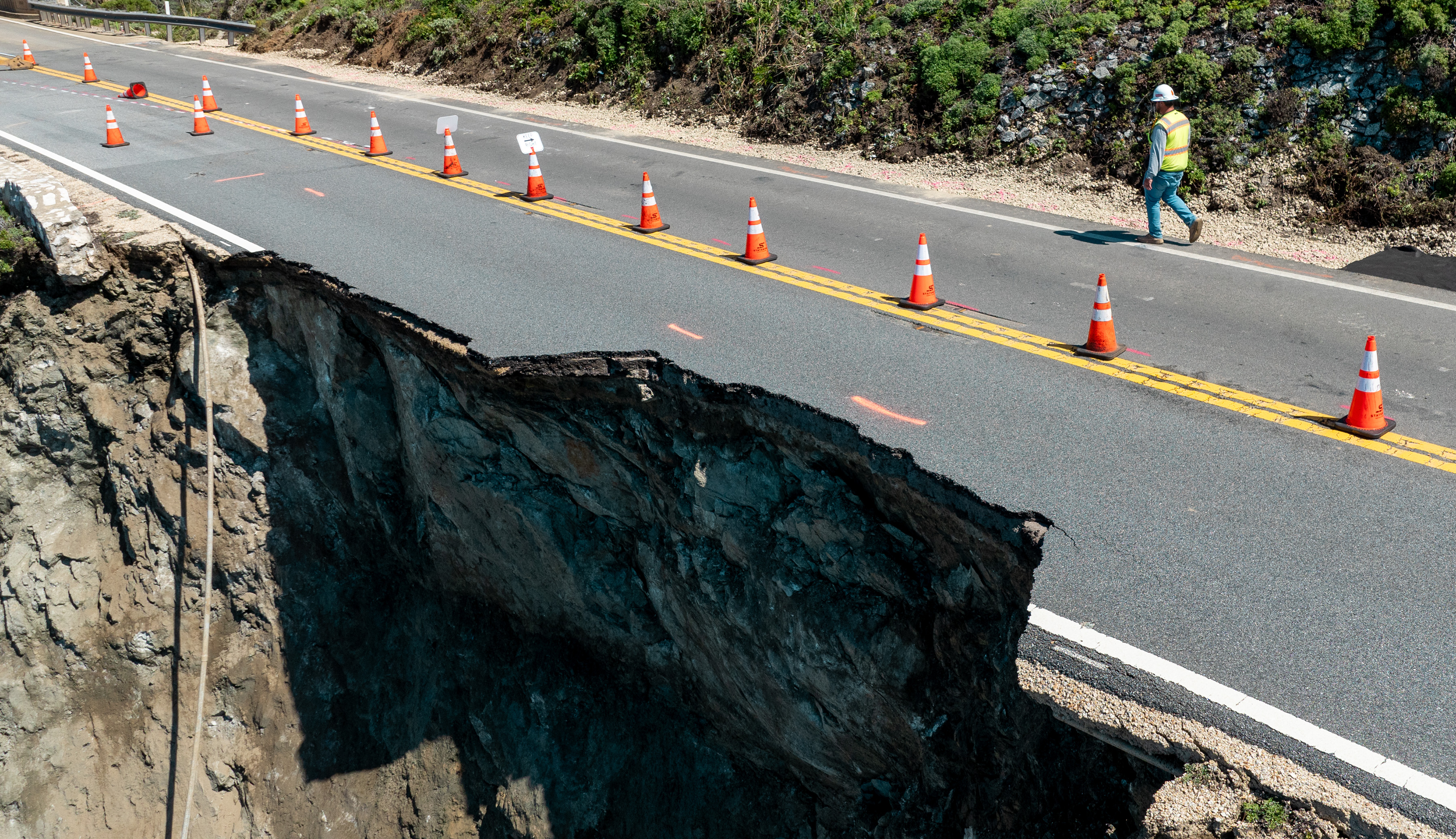 A road has collapsed at the edge, with a deep chasm beside it. Traffic cones mark the area; a person walks by, wearing a safety vest.
