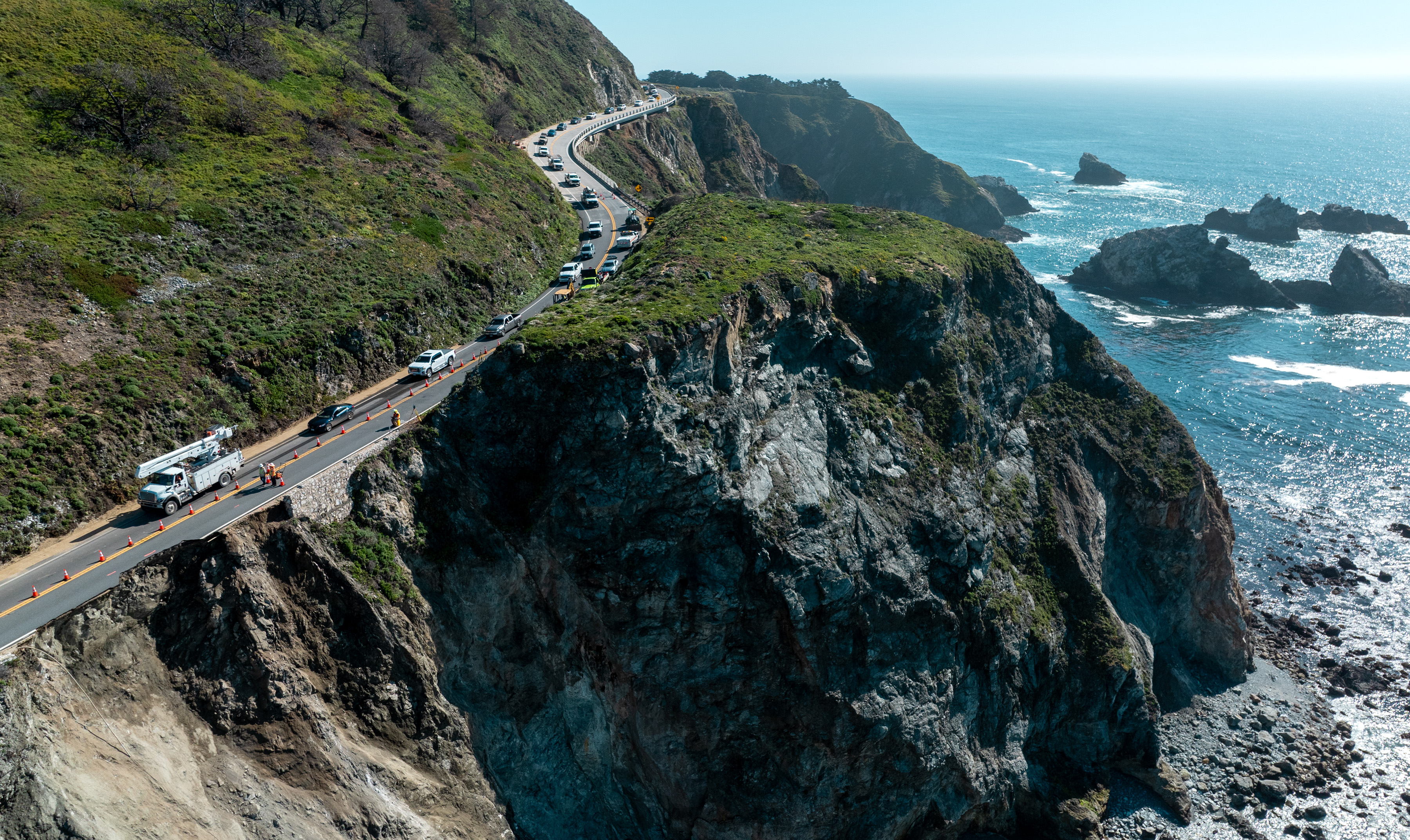 A winding coastal road hugs a steep cliffside above a rocky shoreline, with the ocean in the background.