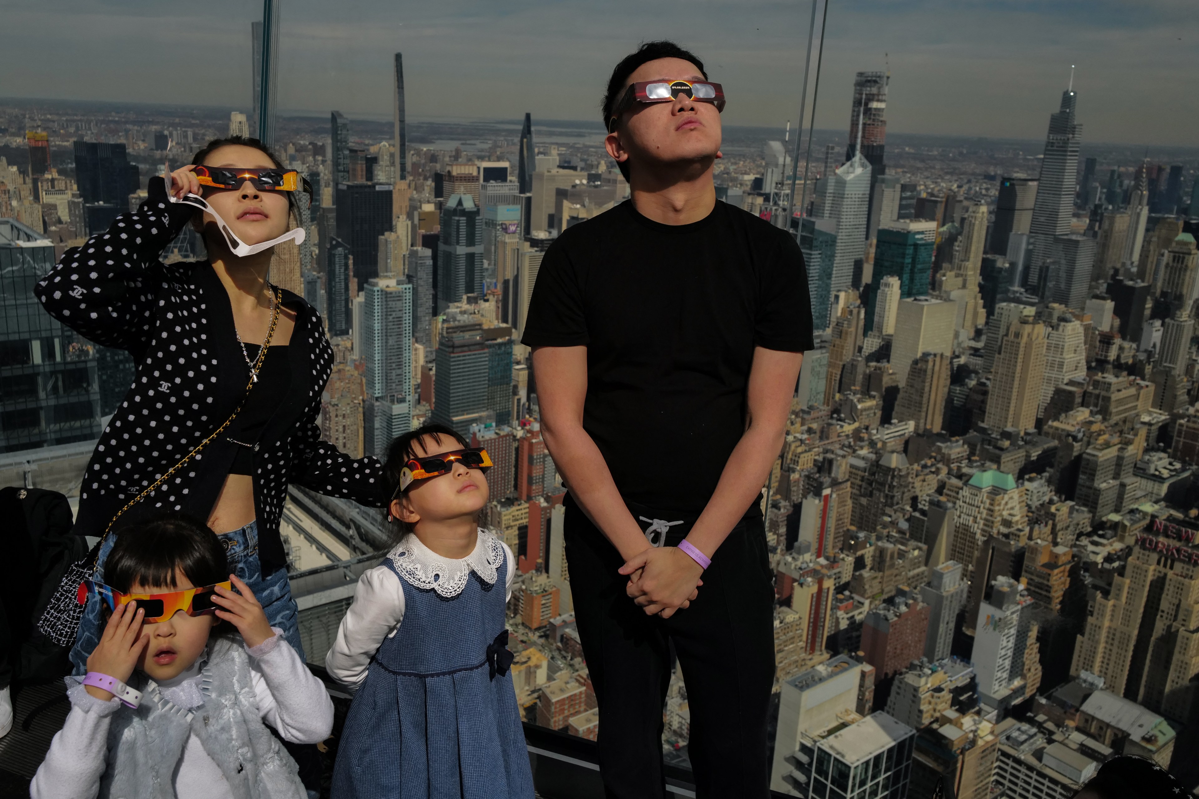 A family of four wearing eclipse glasses stands high above a cityscape, seemingly observing a celestial event.