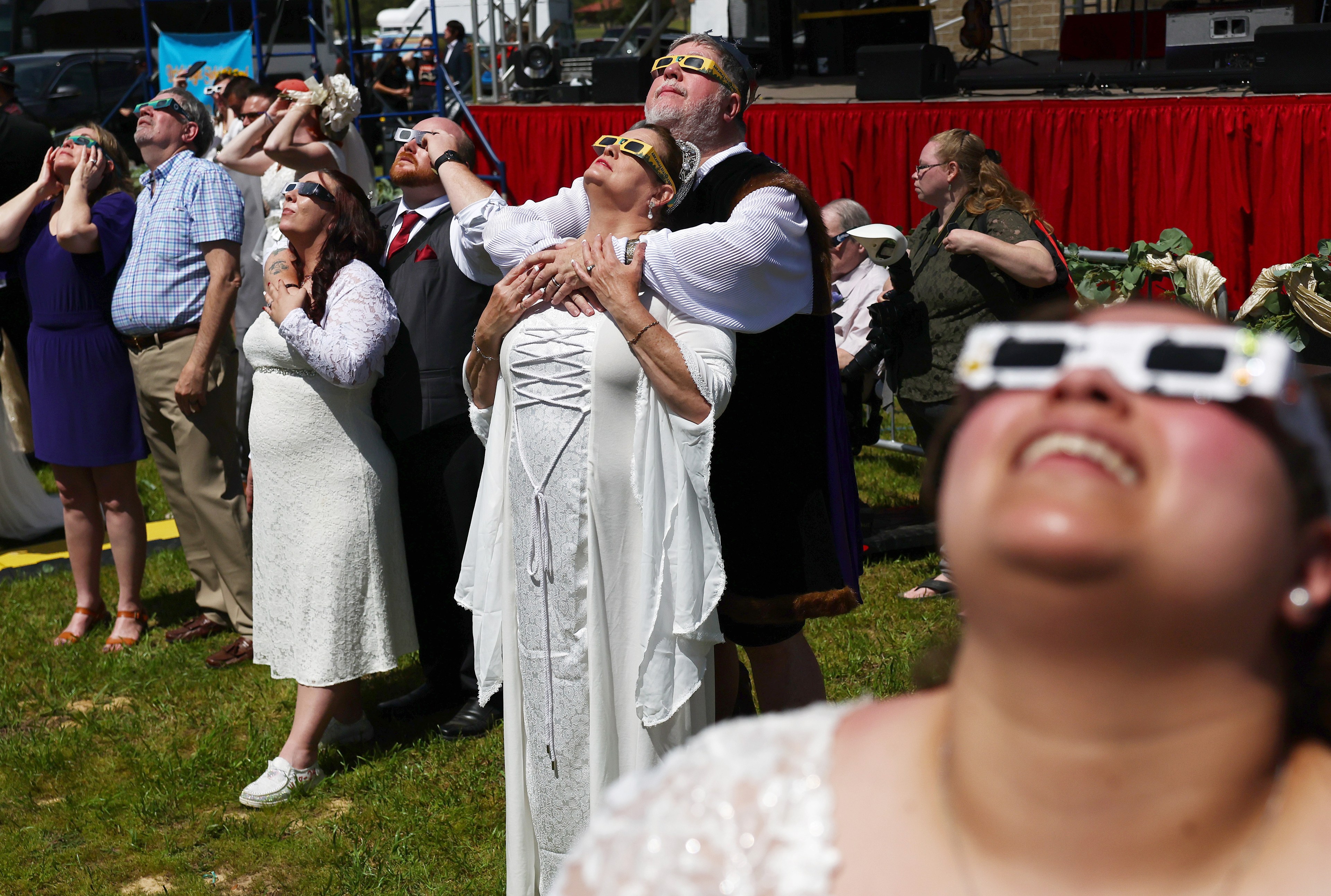 People are wearing special glasses, looking up at the sky; likely viewing an eclipse. A festive atmosphere with a red stage in the background.