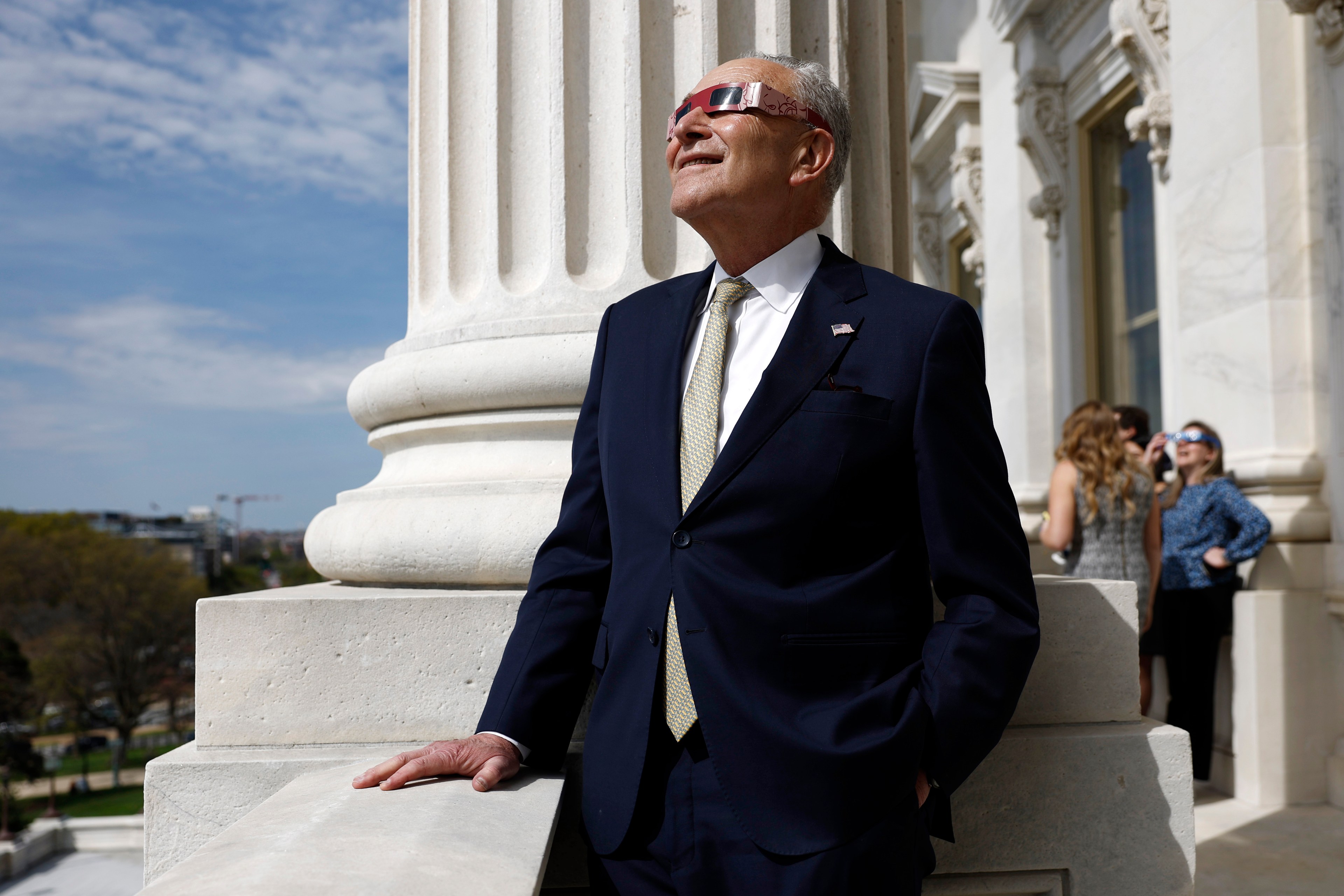 A man in a navy suit stands by a column, wearing eclipse glasses, smiling and looking up. People in background also wear similar glasses.