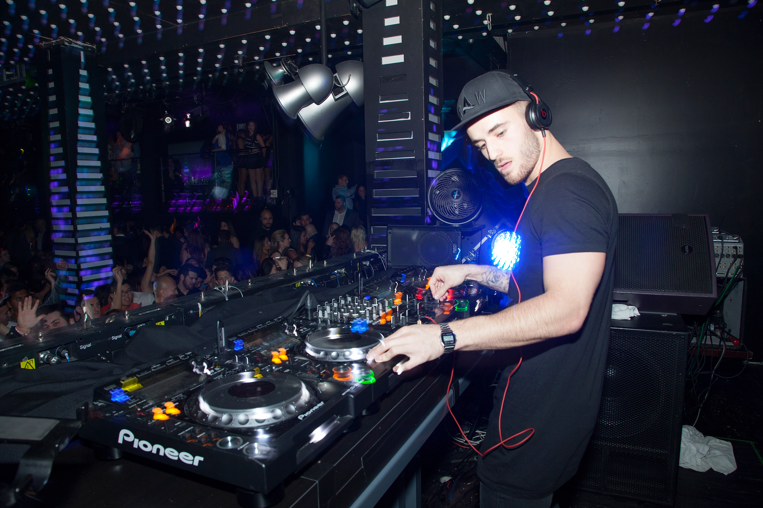 A DJ is mixing music at a club with a crowd in the background, vibrant lights, and a booth with equipment.