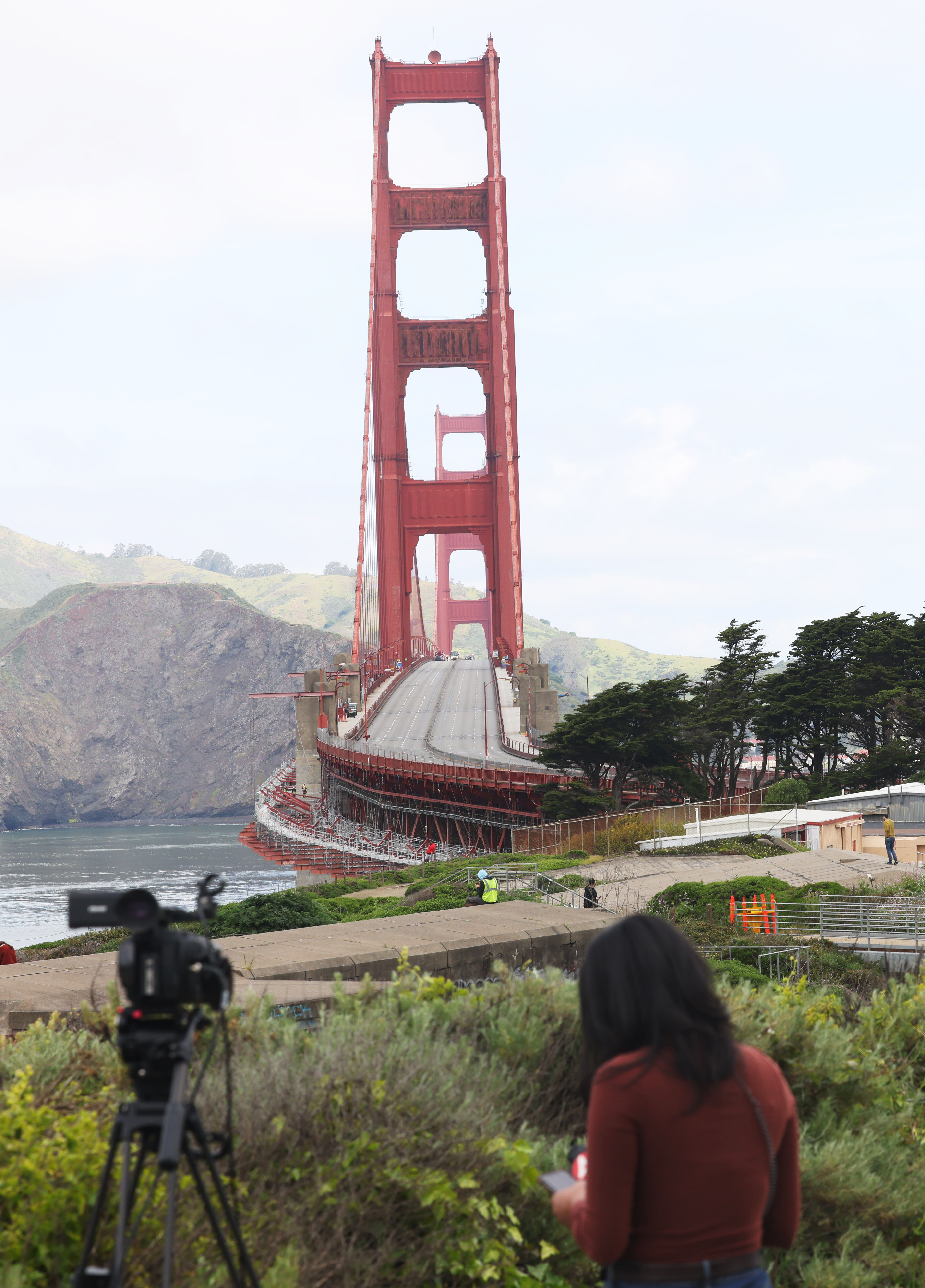 A person is capturing footage of the Golden Gate Bridge; lush greenery and hills surround the iconic red structure.