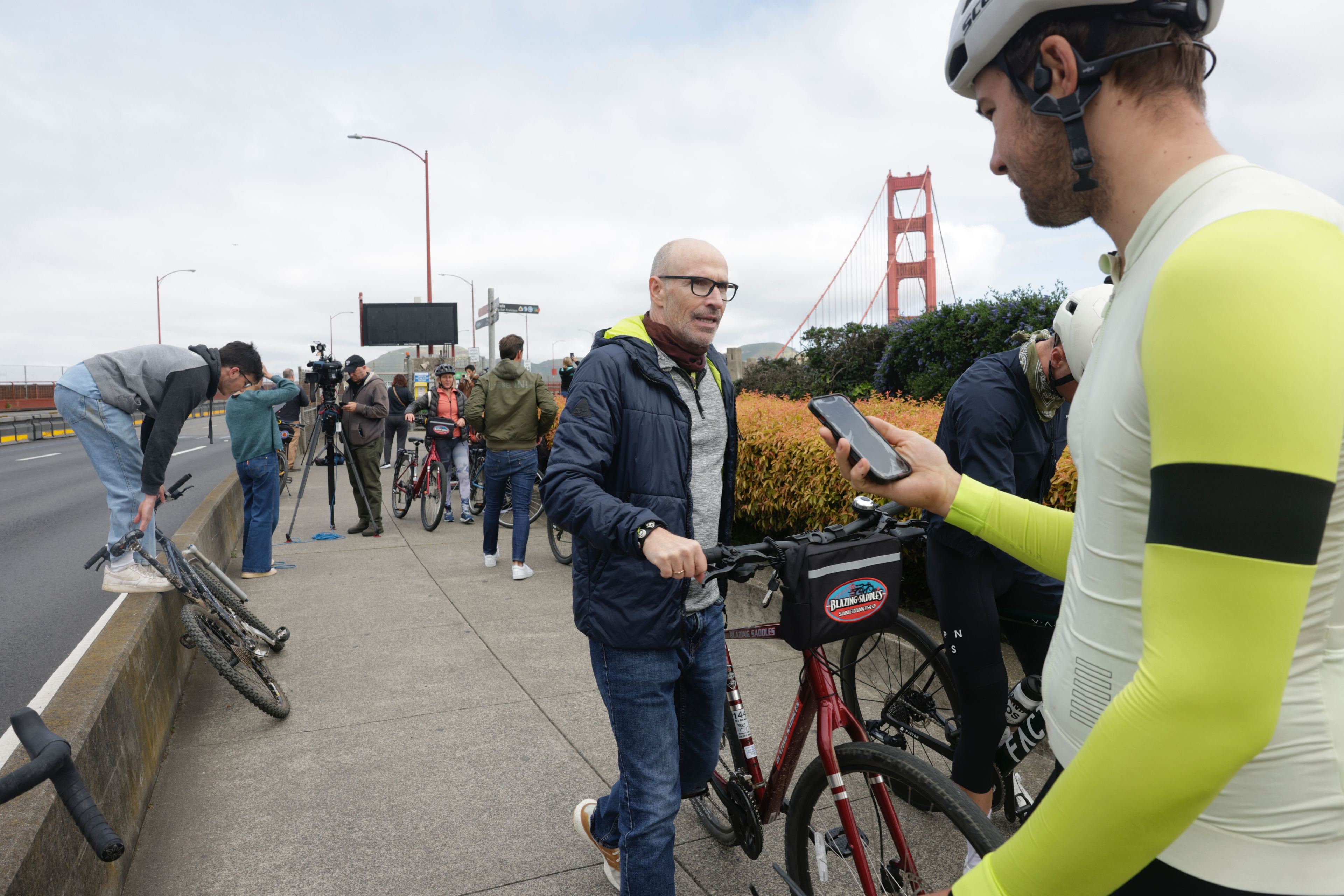 People and cyclists are near the Golden Gate Bridge; one person is looking at their phone.
