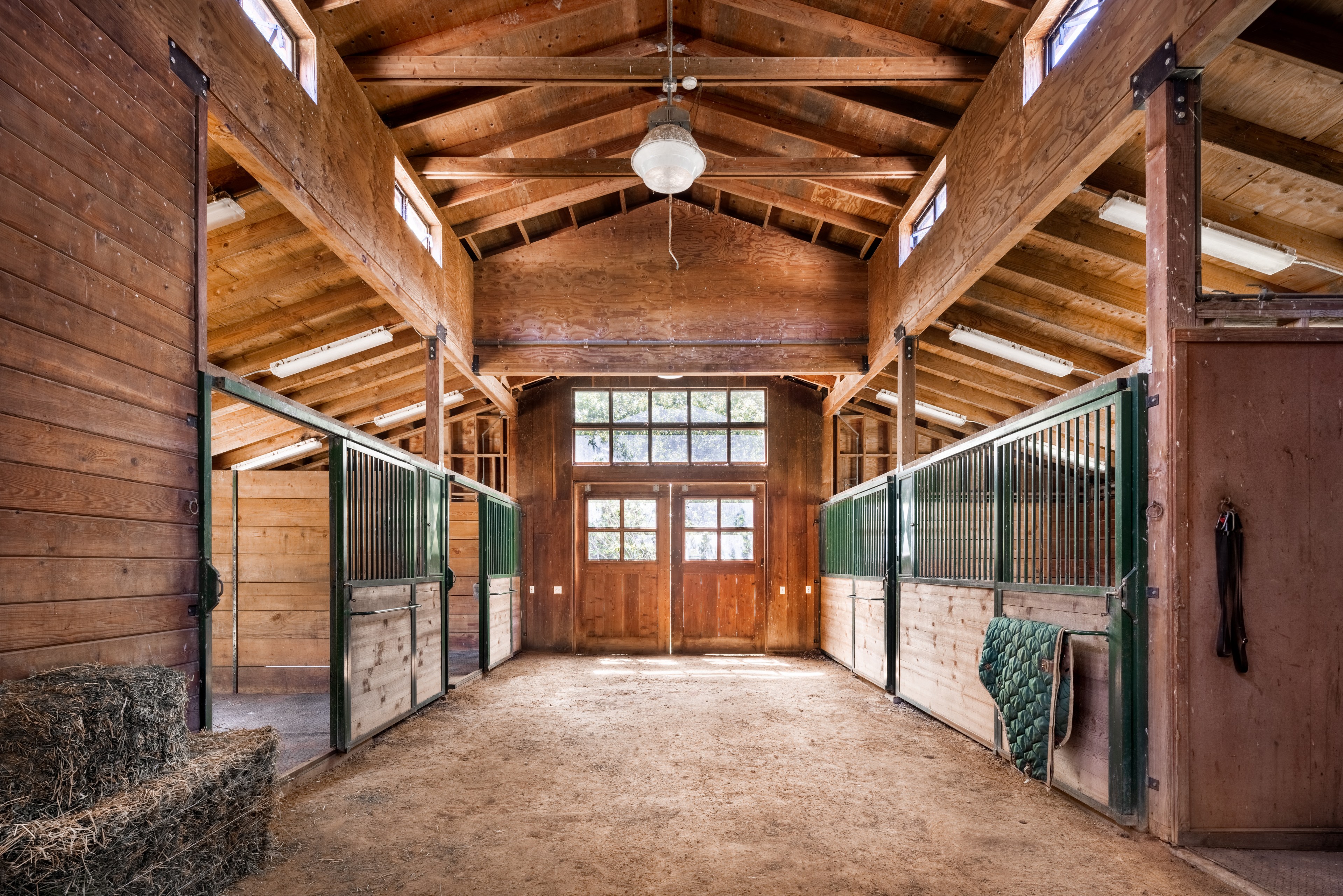 An empty wooden barn with horse stalls, hay bales, and tack hanging on the wall.