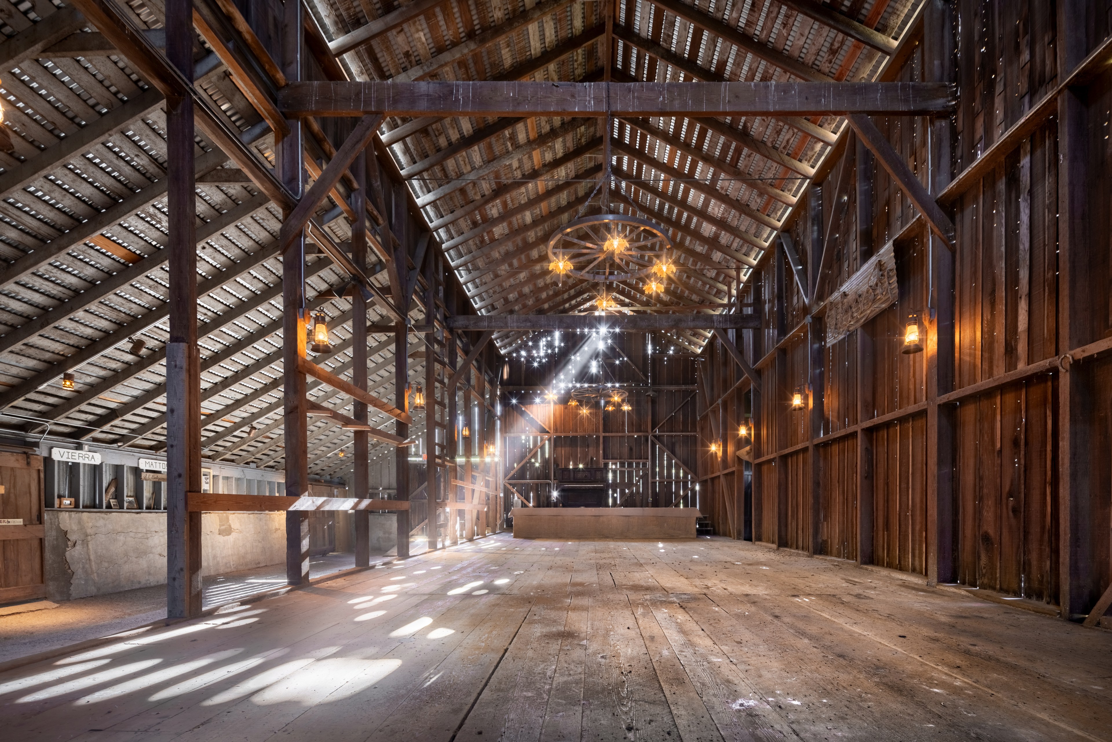Inside an empty rustic barn with wooden beams, hanging lights, and sunbeams filtering through the walls.