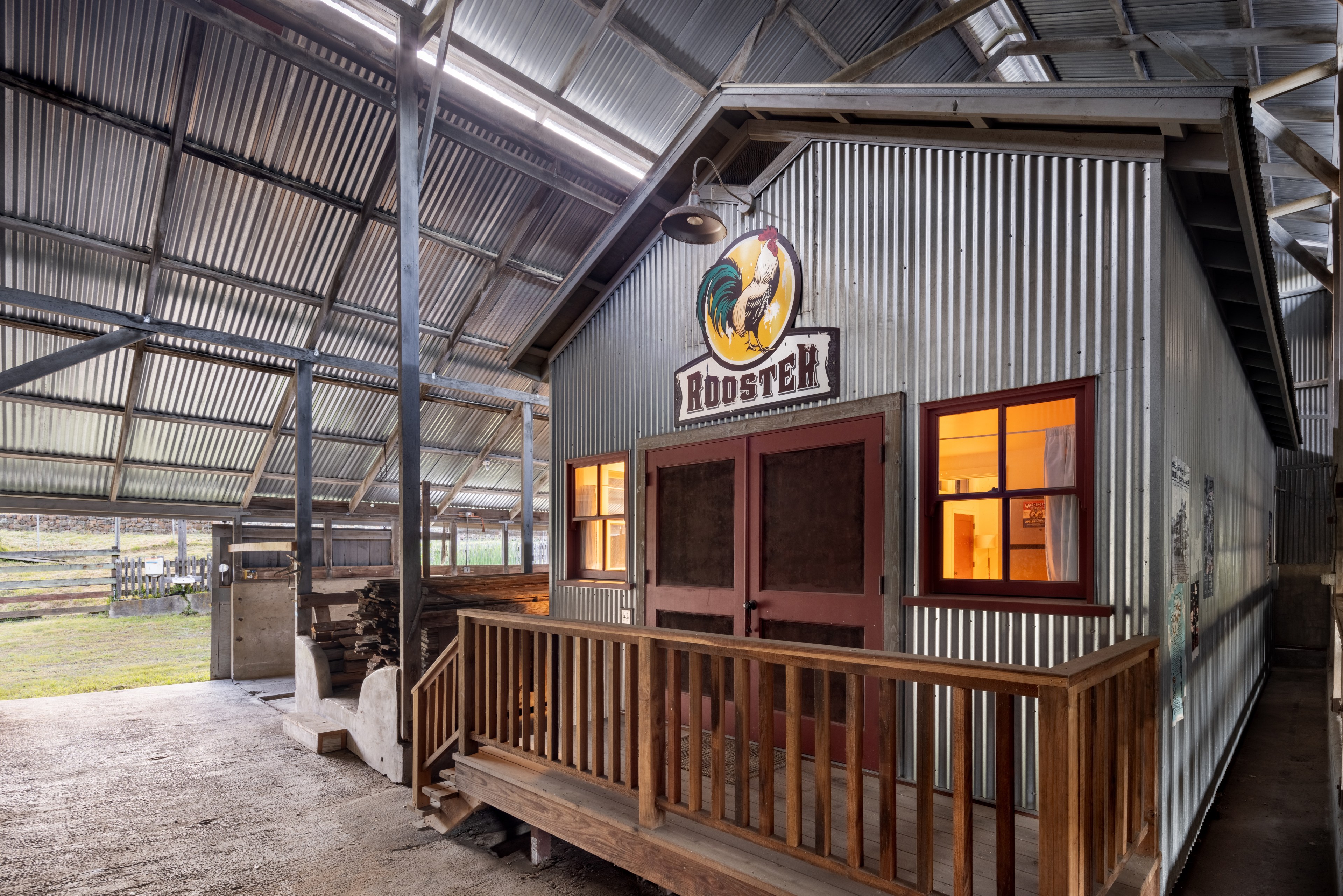 A rustic barn interior with a porch, corrugated metal walls, and a &quot;Rooster&quot; sign. Warm light pours from a window.