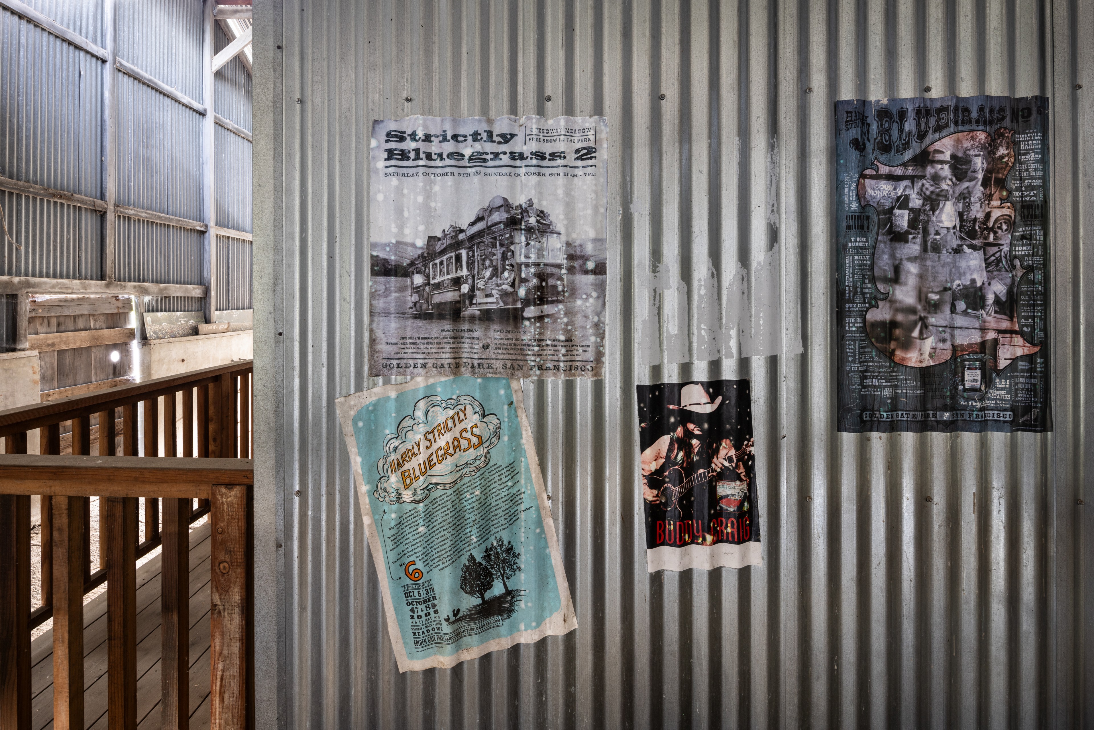 The image shows a corrugated metal wall with four vintage music posters, each with unique designs, in a rustic setting with wooden railings on the left.