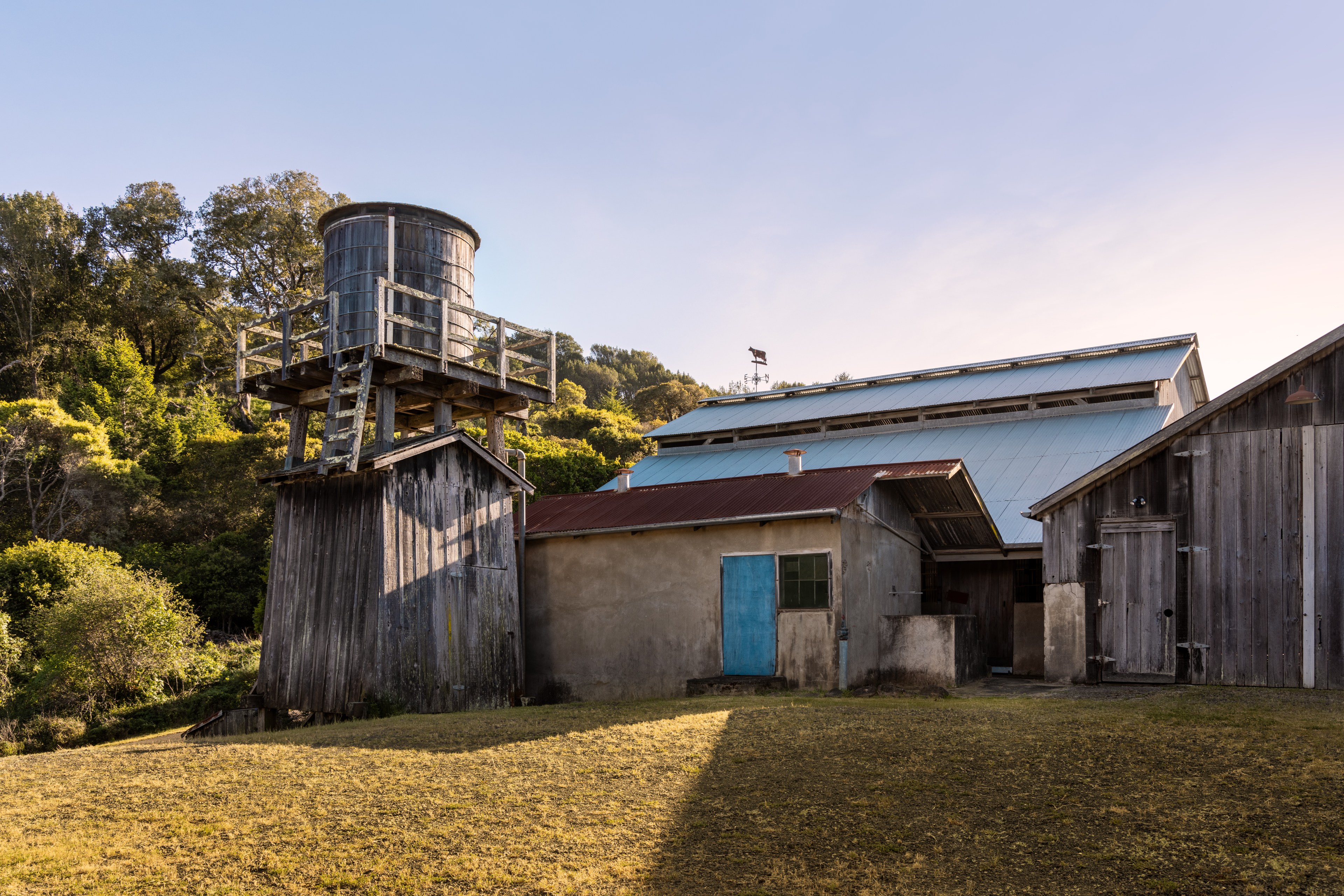 The image shows a rustic wooden water tower with a ladder, alongside several old buildings with metal and wooden roofs, set against a backdrop of trees and clear sky.