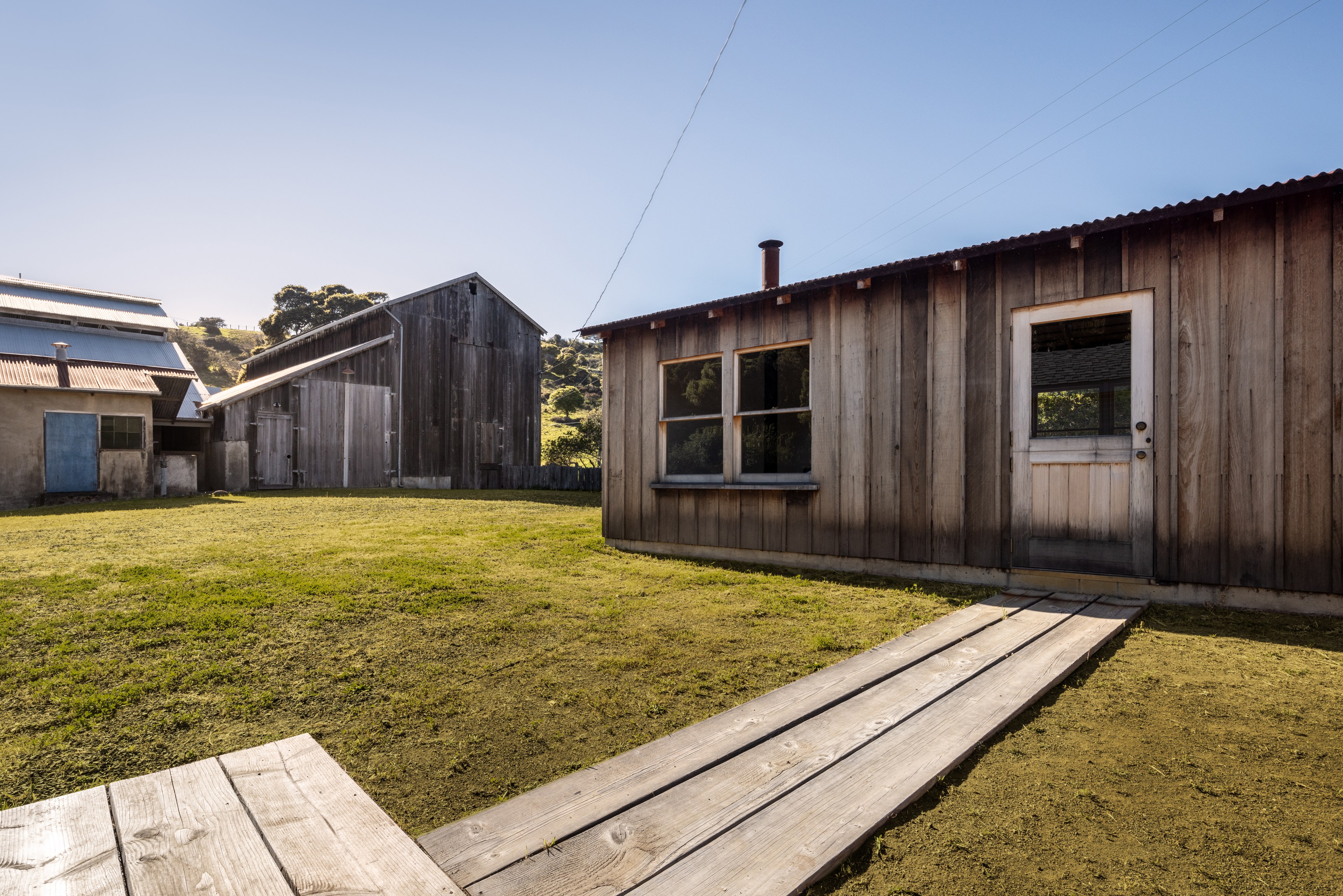 The image shows a sunny outdoor scene with rustic wooden buildings, a grassy area, and a wooden walkway. The structures have weathered wood and corrugated metal roofs.