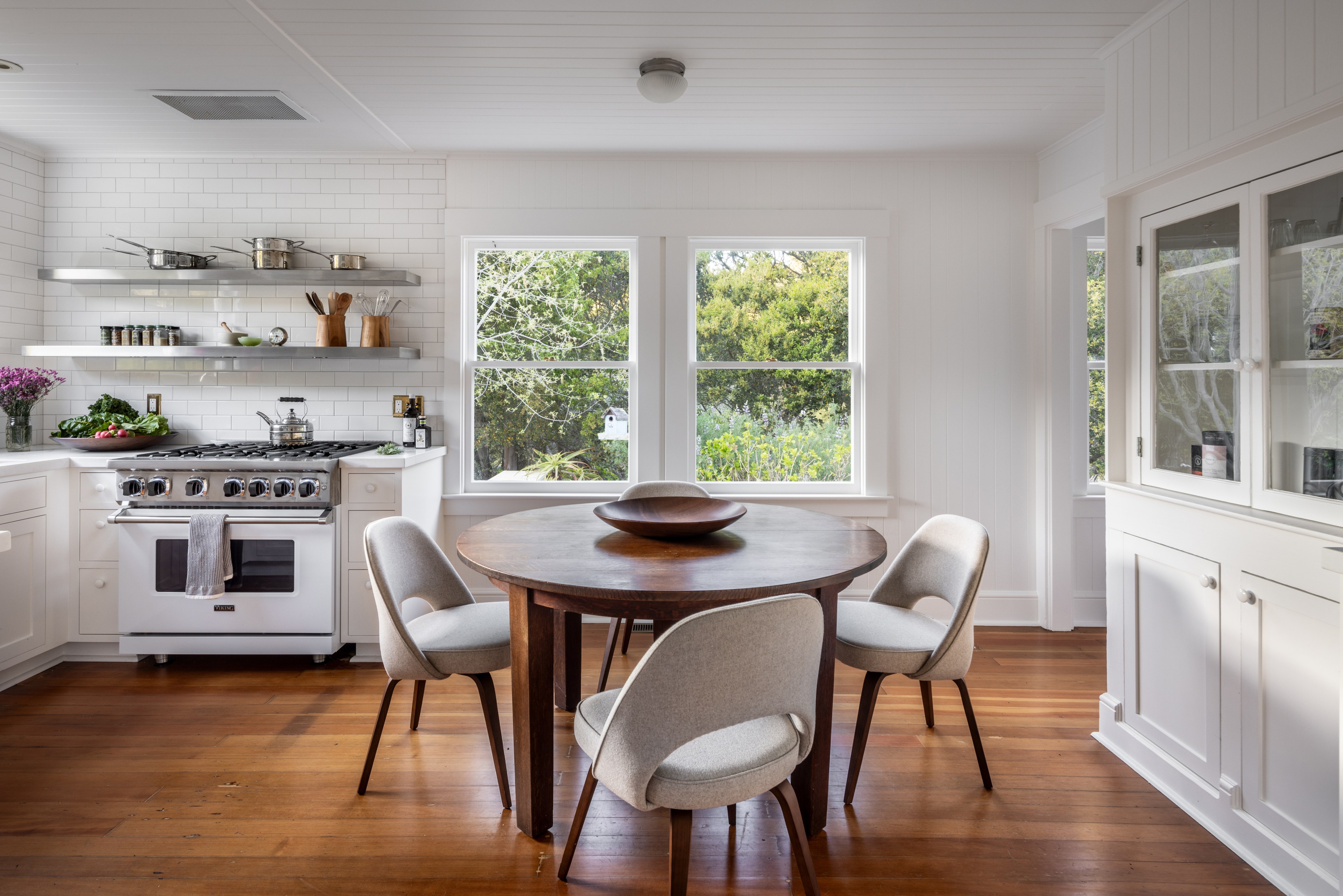 A bright kitchen with white cabinets, a round table, chairs, and a view of greenery outside the window.