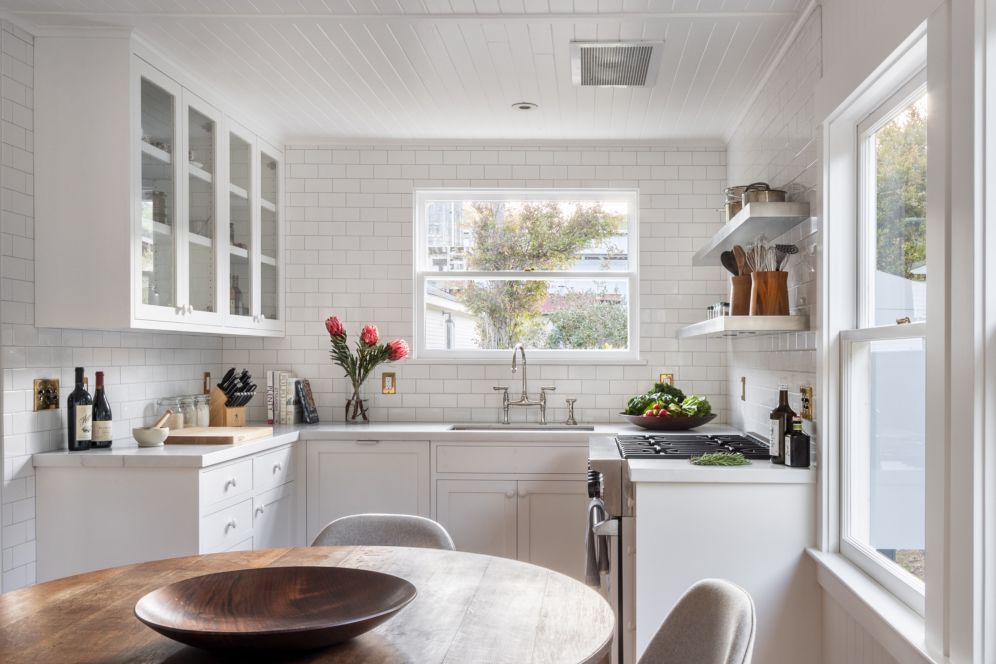 A bright kitchen with white cabinets and subway tiles, featuring a window, wooden dining table, and countertop with kitchen essentials and fresh produce.