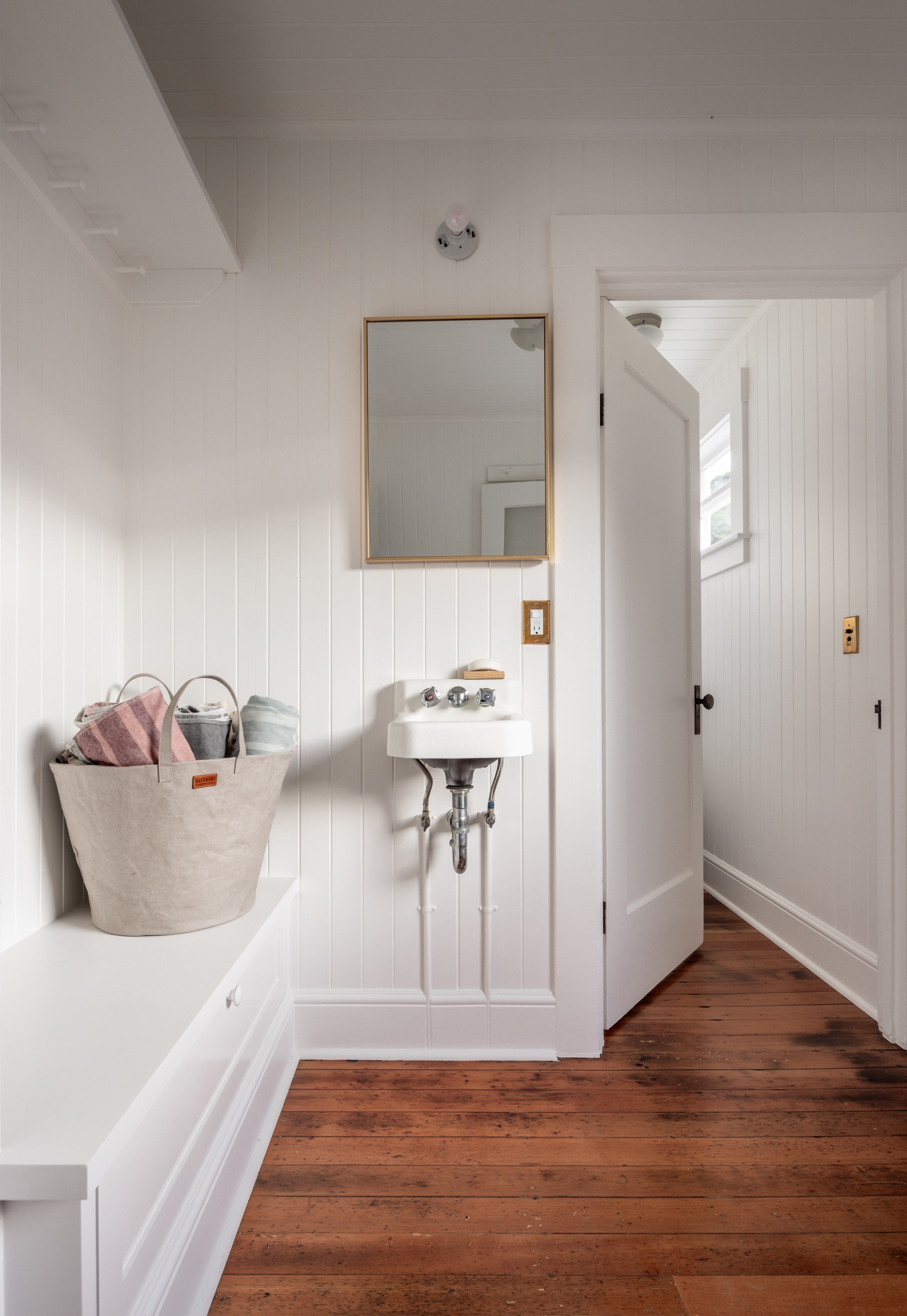 The image shows a small, white-painted bathroom with wooden floors, a wall-mounted sink, a mirror above it, and a basket with towels on a bench.