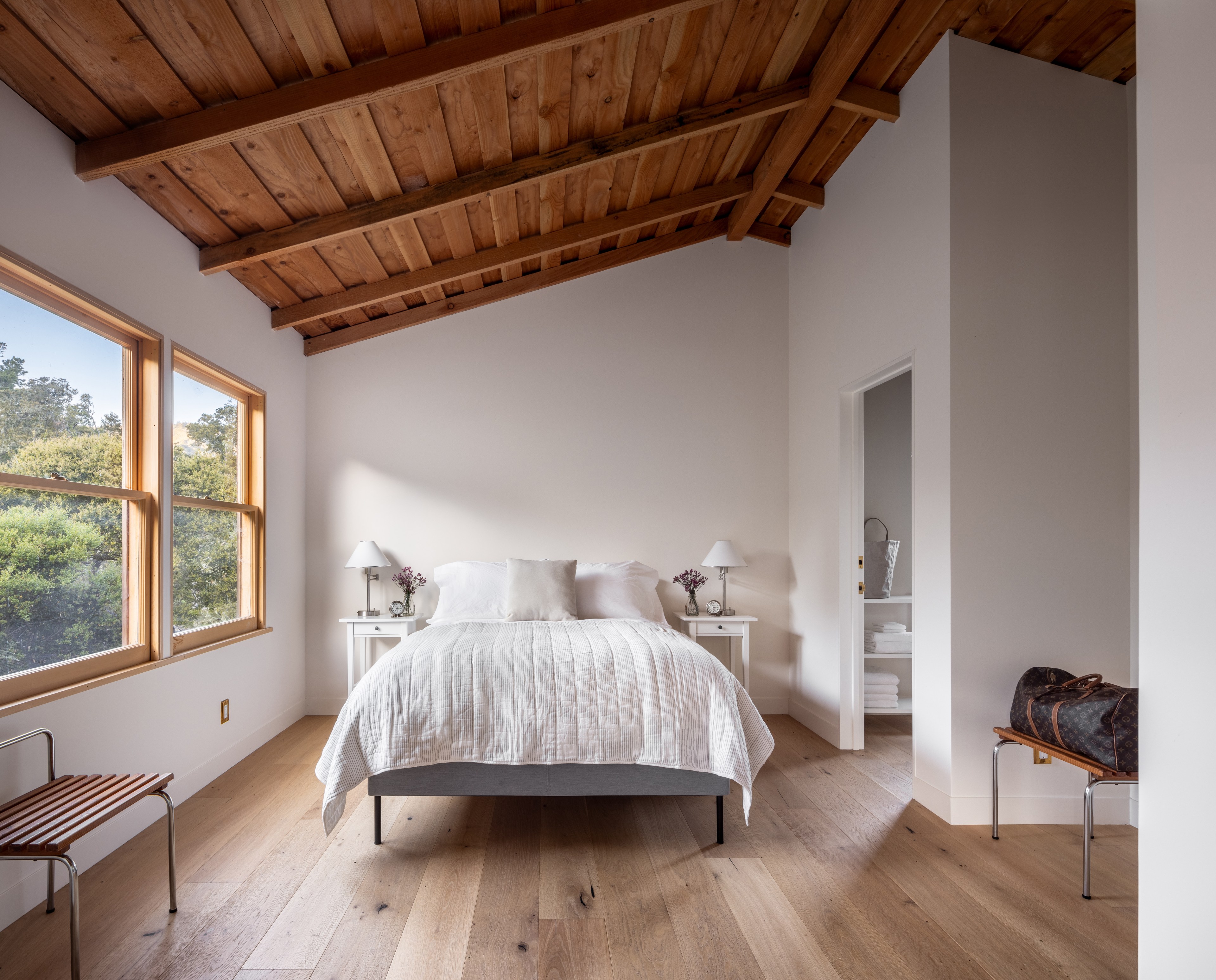 A serene bedroom with a wood beam ceiling, wooden floors, a double bed, two nightstands with lamps, and windows overlooking greenery.
