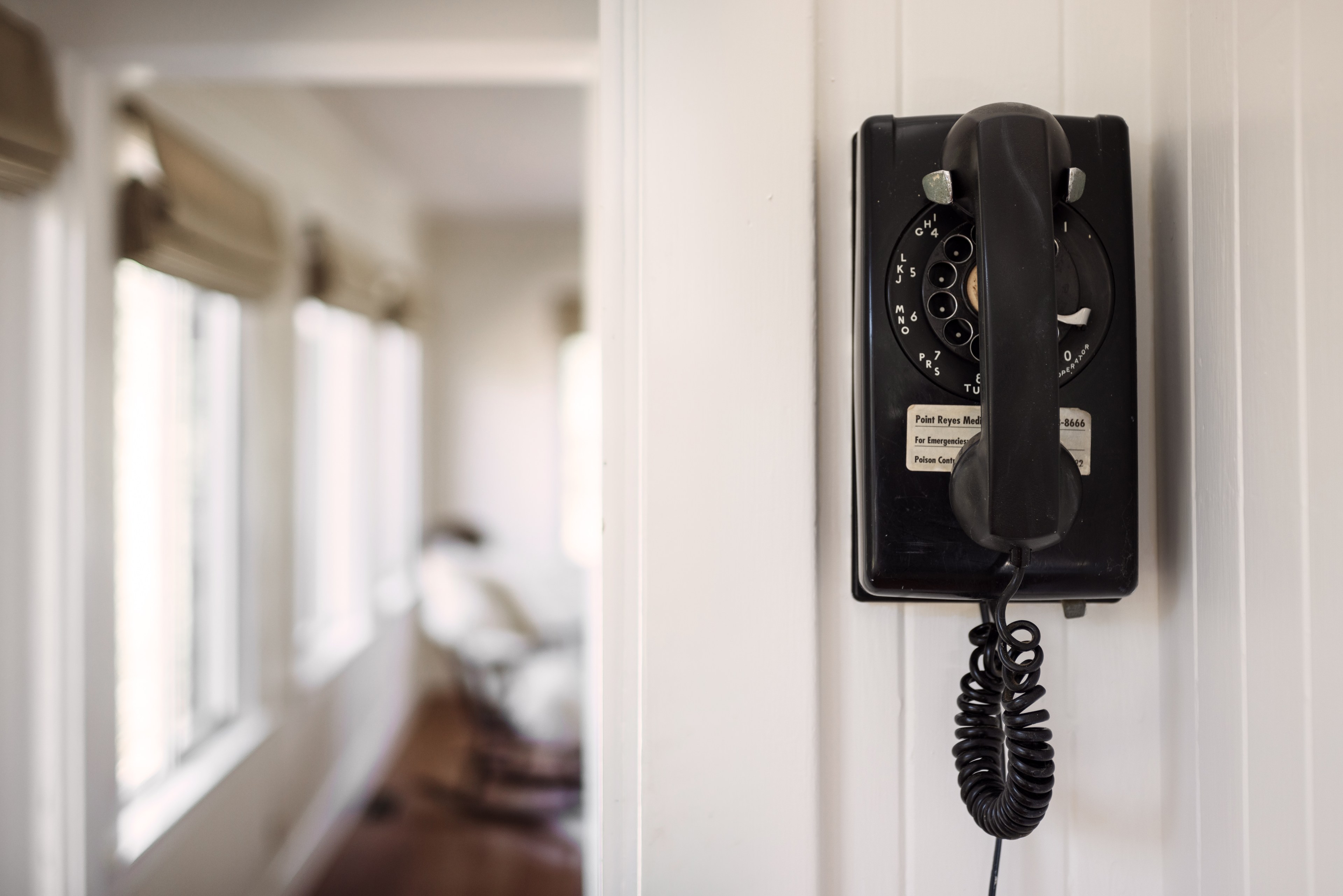 A black rotary wall phone is mounted on a white paneled wall. Its coiled cord hangs below, with a blurred room and windows visible in the background.